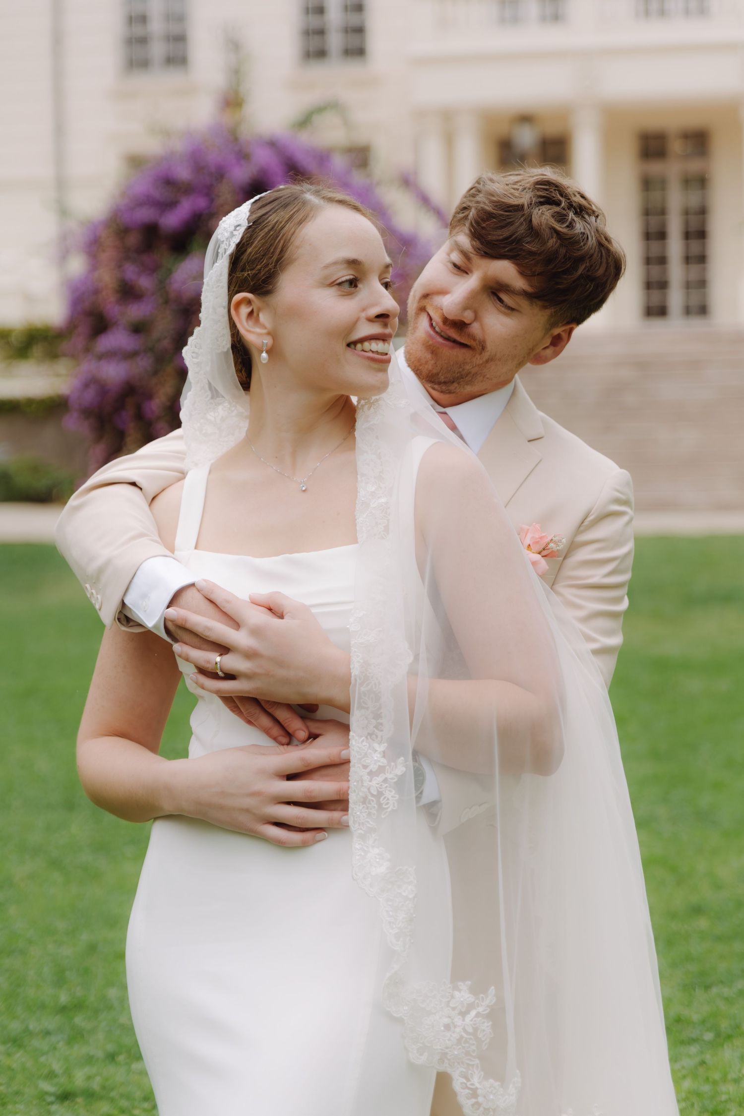Groom in beige suit embraces bride from behind on garden lawn before elegant manor with purple flowering shrubs.