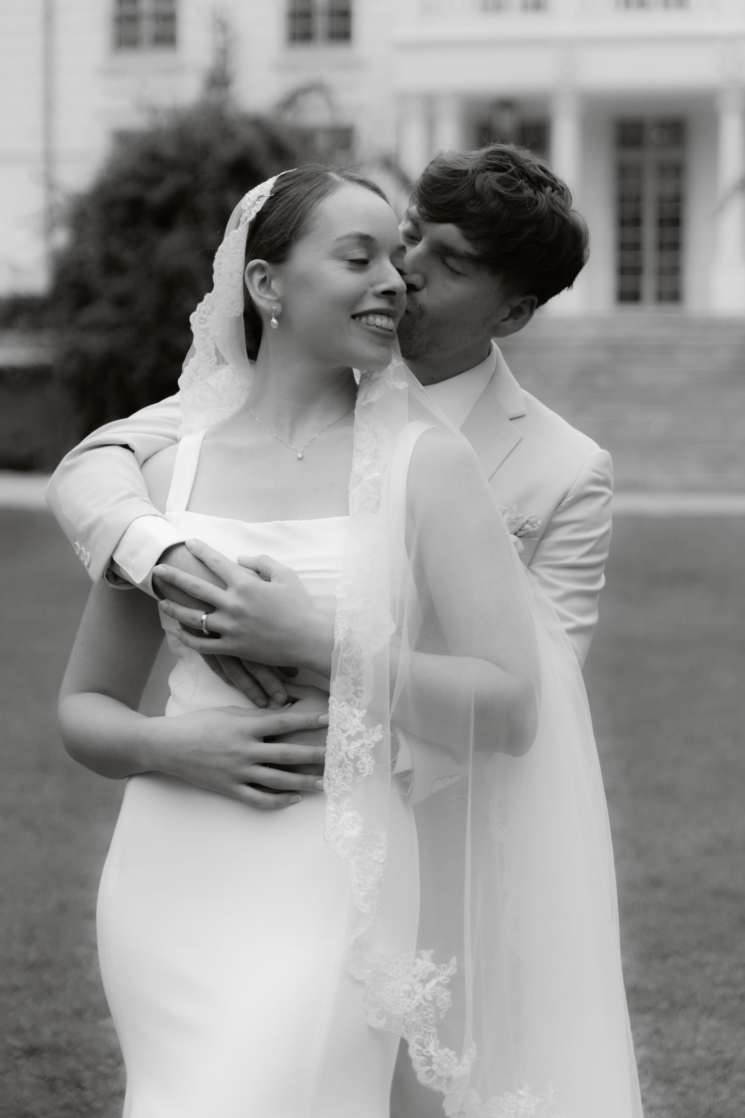 Black and white photo of groom kissing smiling bride in lace veil on lawn before grand wedding venue.