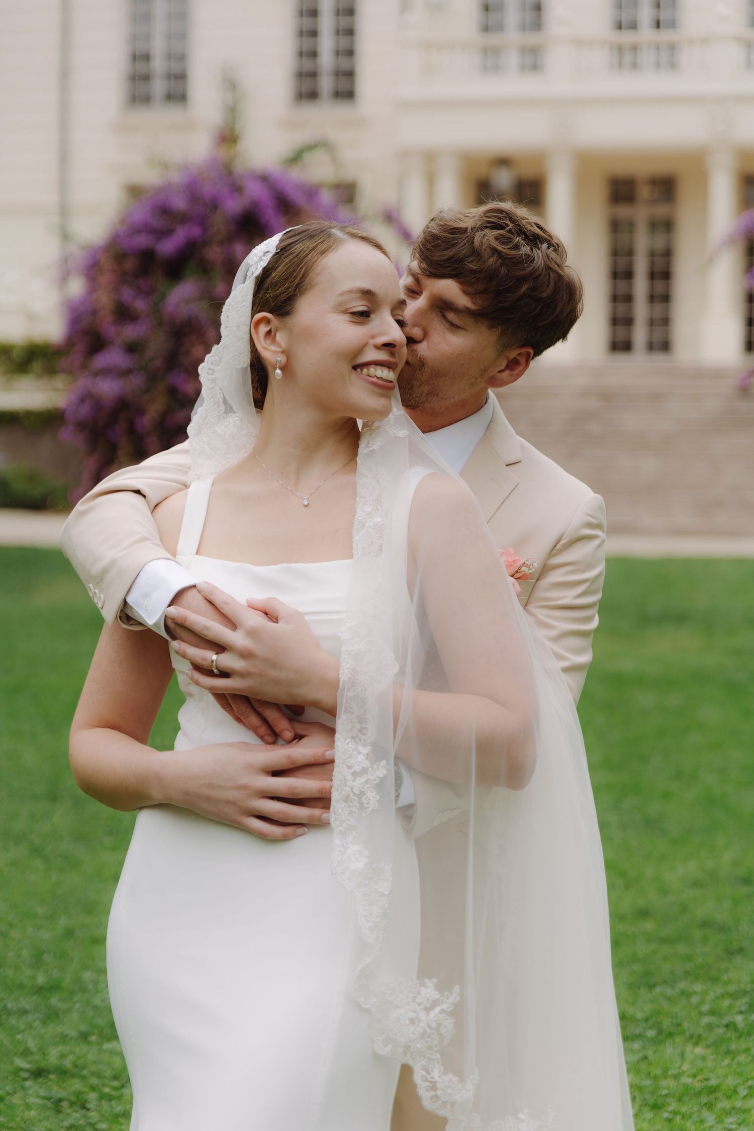 Groom tenderly kisses laughing bride in lace veil and white gown on green lawn before elegant manor.