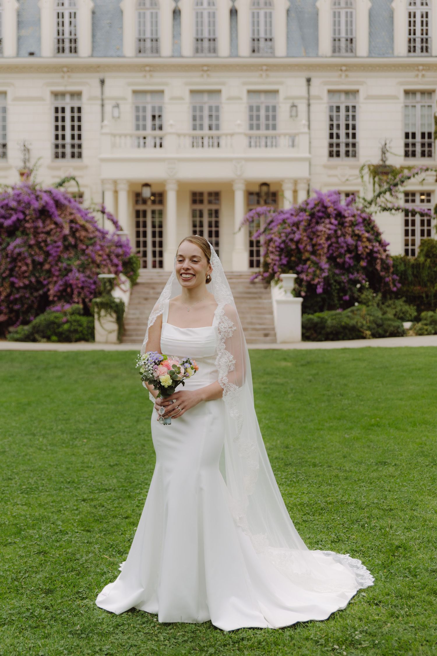 Bride in white gown with lace veil holds colorful bouquet on lawn before elegant mansion with purple flowers.