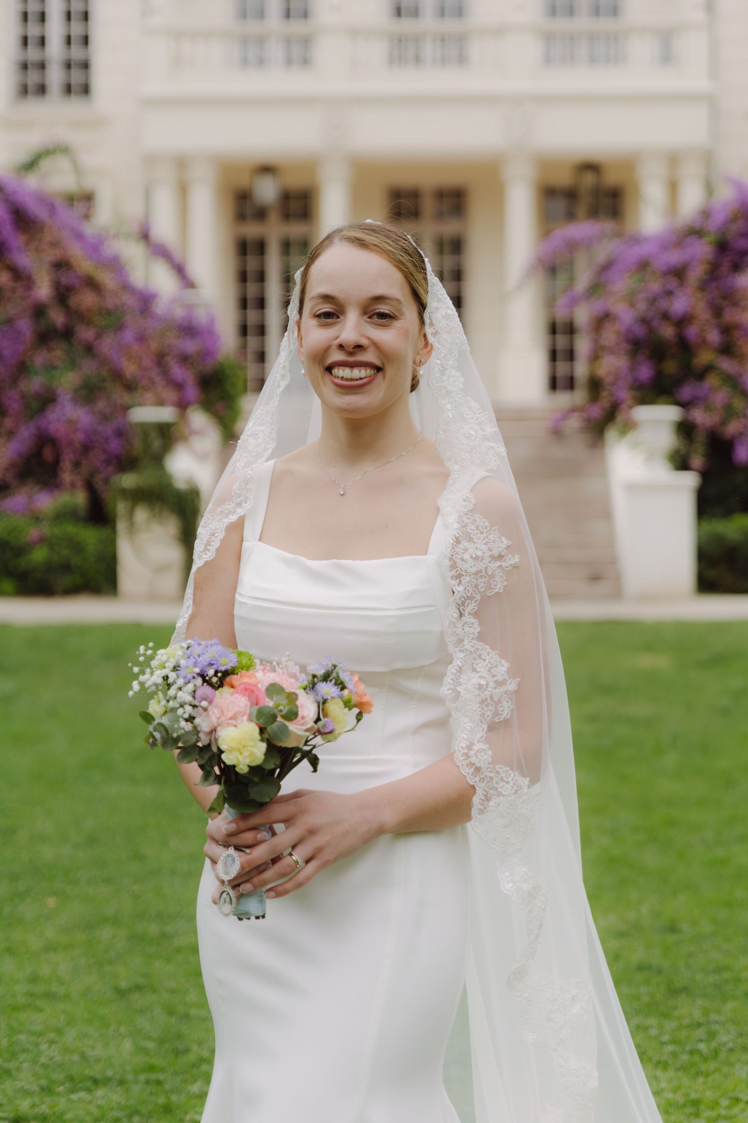 Smiling bride in fitted white dress and lace cathedral veil holds pastel bouquet before grand estate.