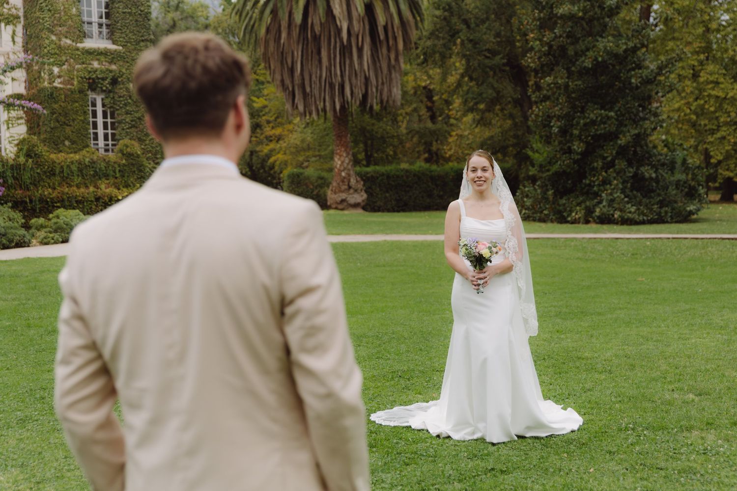 Groom in beige suit faces bride in white gown with veil on lush green lawn at chateau venue.