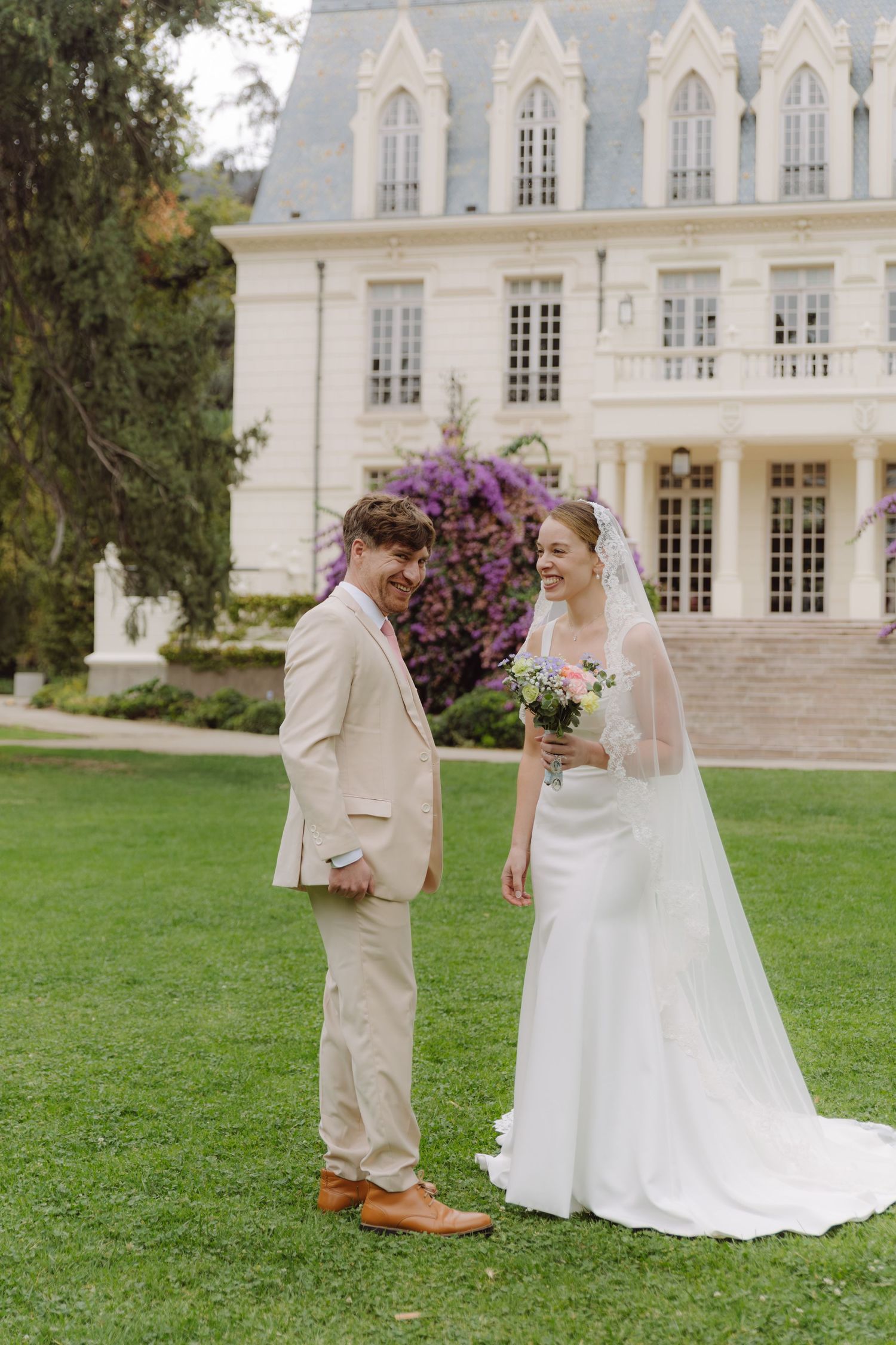 Bride and groom share a joyful moment on green lawn before a grand white chateau with purple blooms.