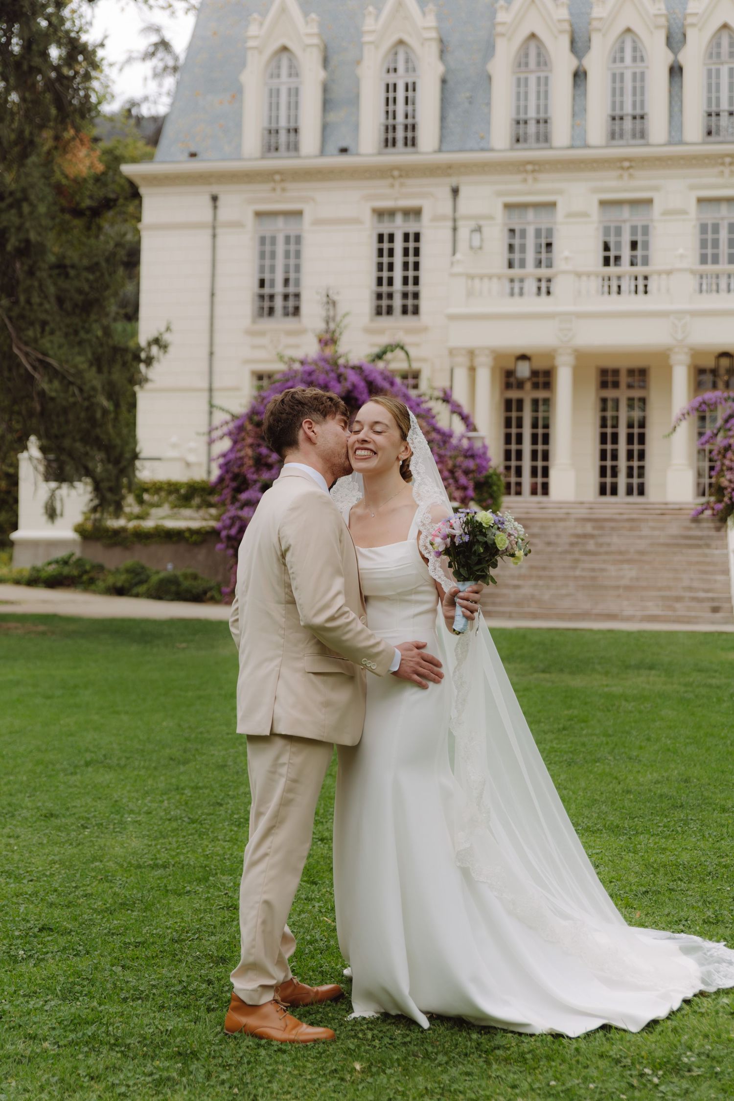 Groom in tan suit whispers to smiling bride in white gown holding bouquet before elegant chateau.