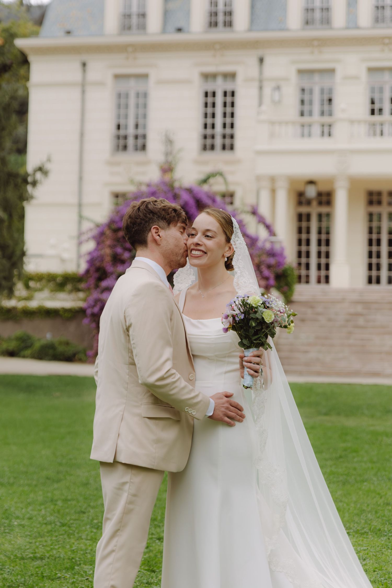 Bride and groom share an intimate moment on lawn before white chateau adorned with purple wisteria.