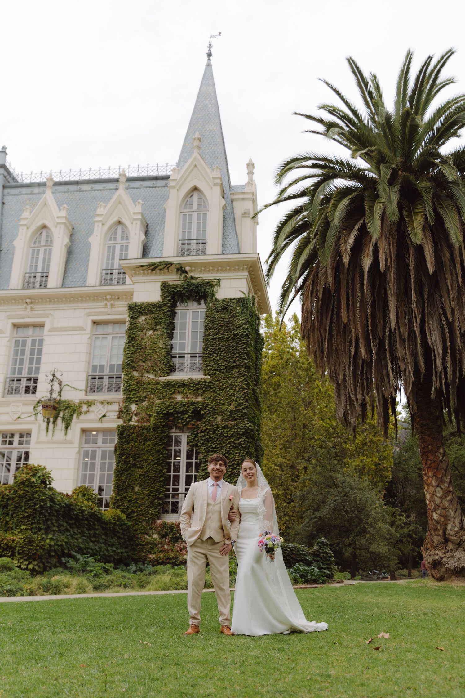 Bride and groom walk hand in hand before an ivy-covered Gothic chateau with tall palm tree.