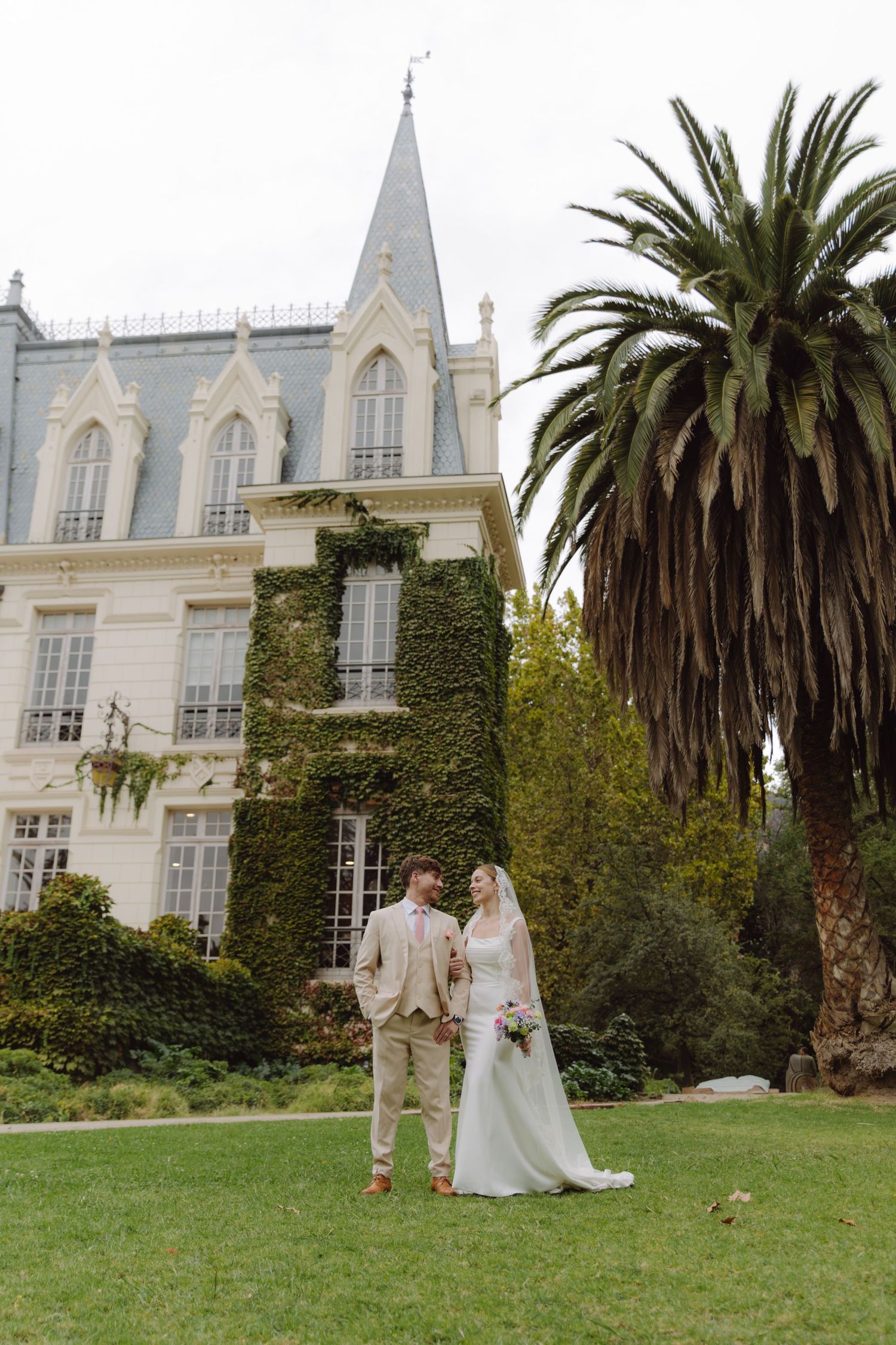 Newlyweds stand together on green lawn before ivy-clad Gothic chateau with towering palm tree.