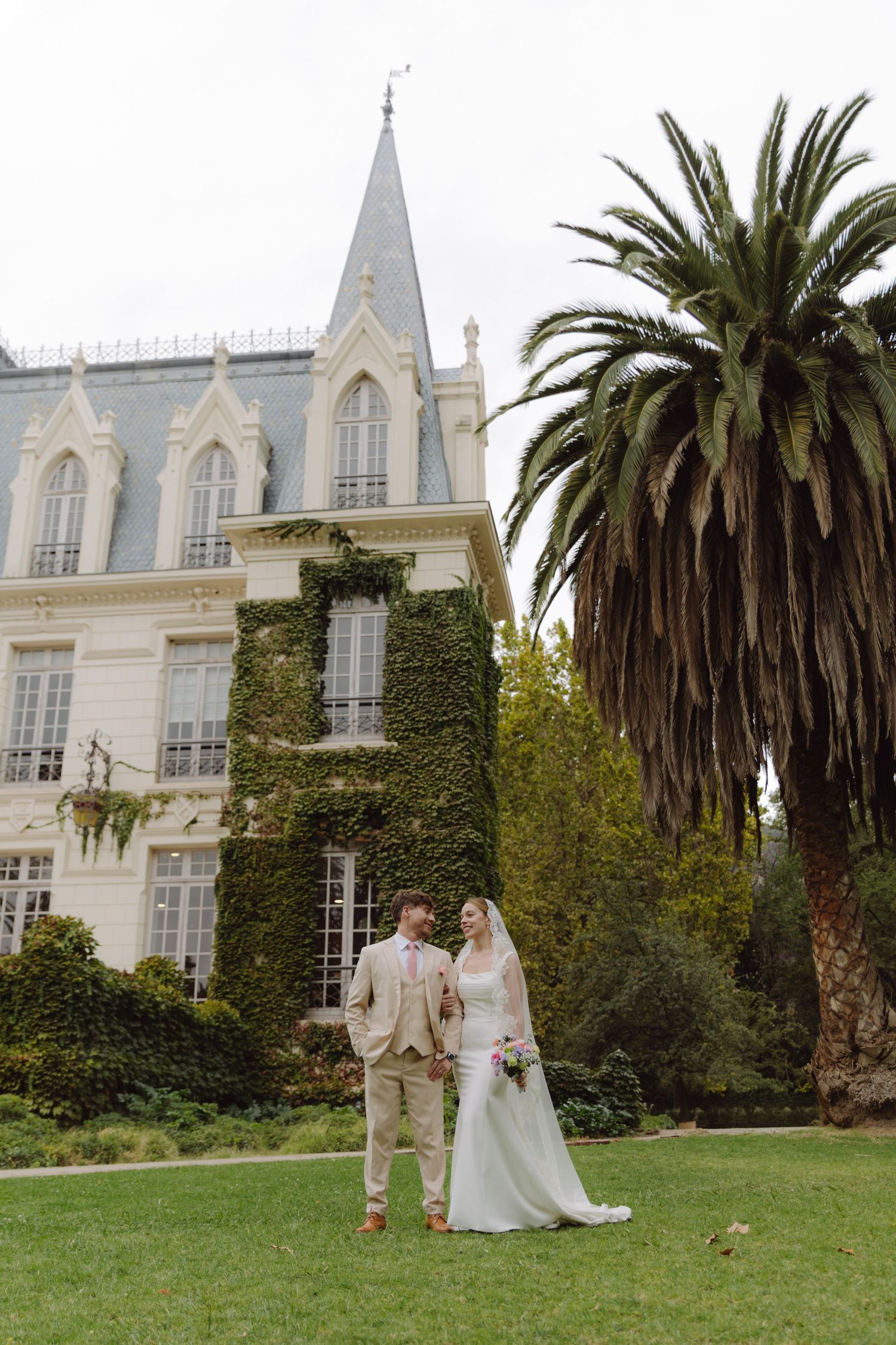 Bride in white gown and groom in beige suit pose before ivy-covered chateau with Gothic spire and palm.