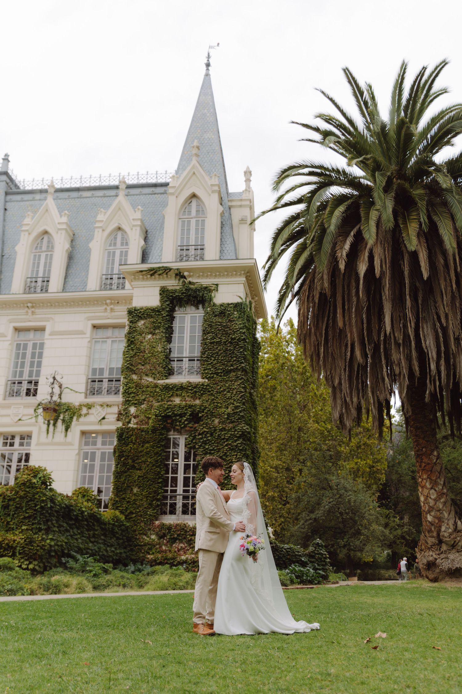 Bride and groom face each other tenderly before ivy-covered Gothic chateau with palm tree backdrop.