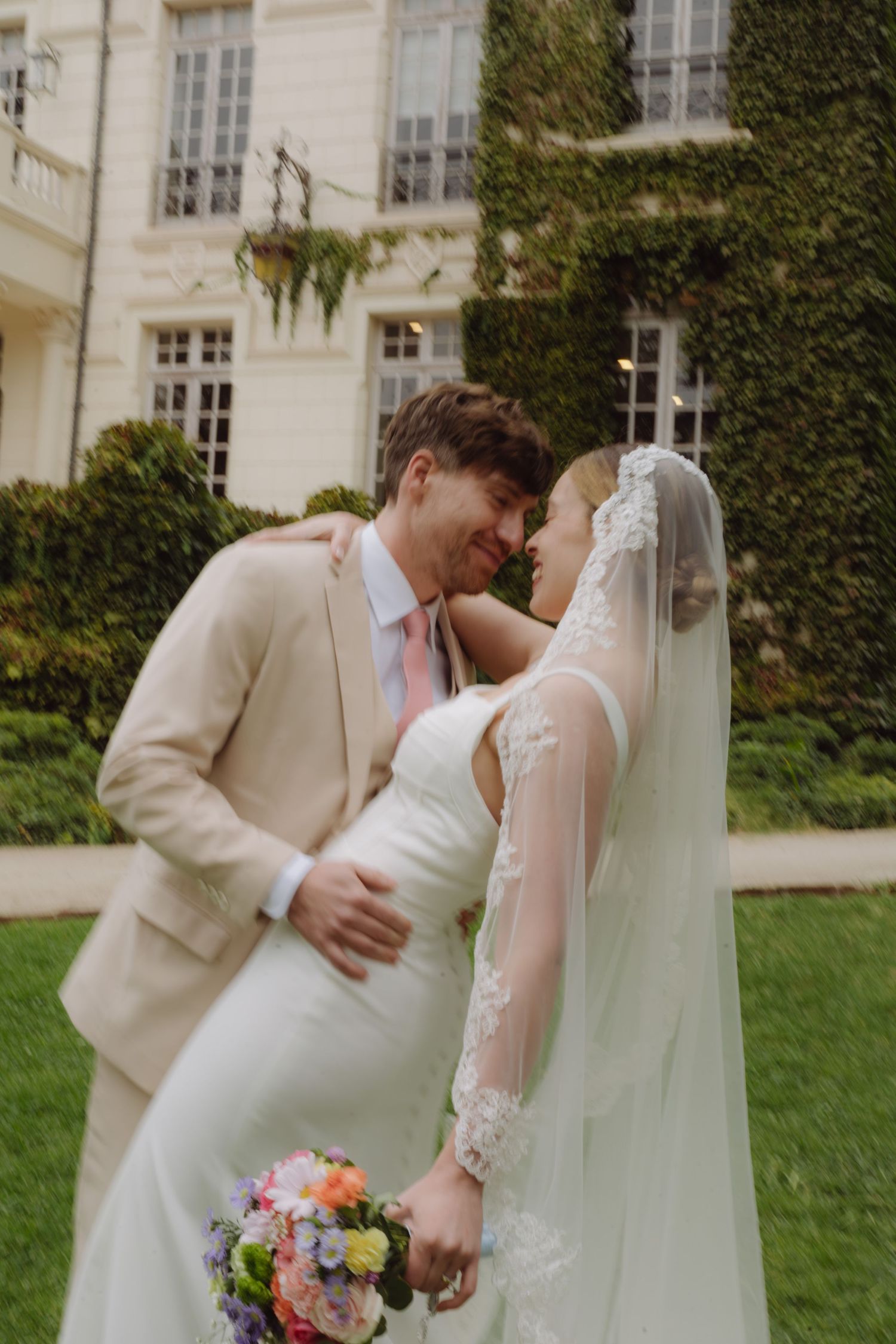 Bride and groom share a romantic moment in garden, groom in beige suit, bride in white gown with lace veil holding colorful bouquet.