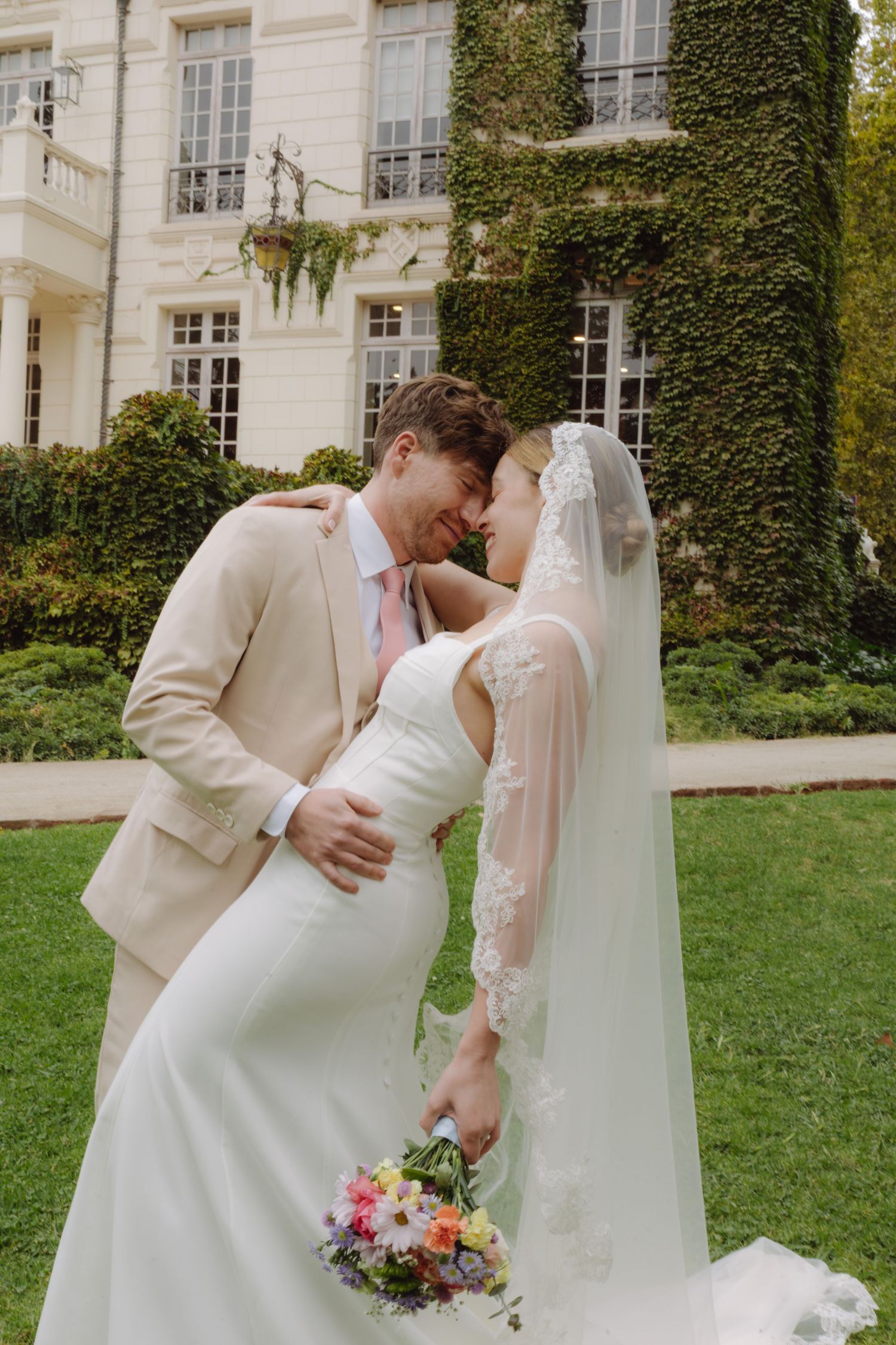 Wedding couple nearly kissing outdoors, groom in tan suit with pink tie, bride in fitted white dress with lace veil and floral bouquet.