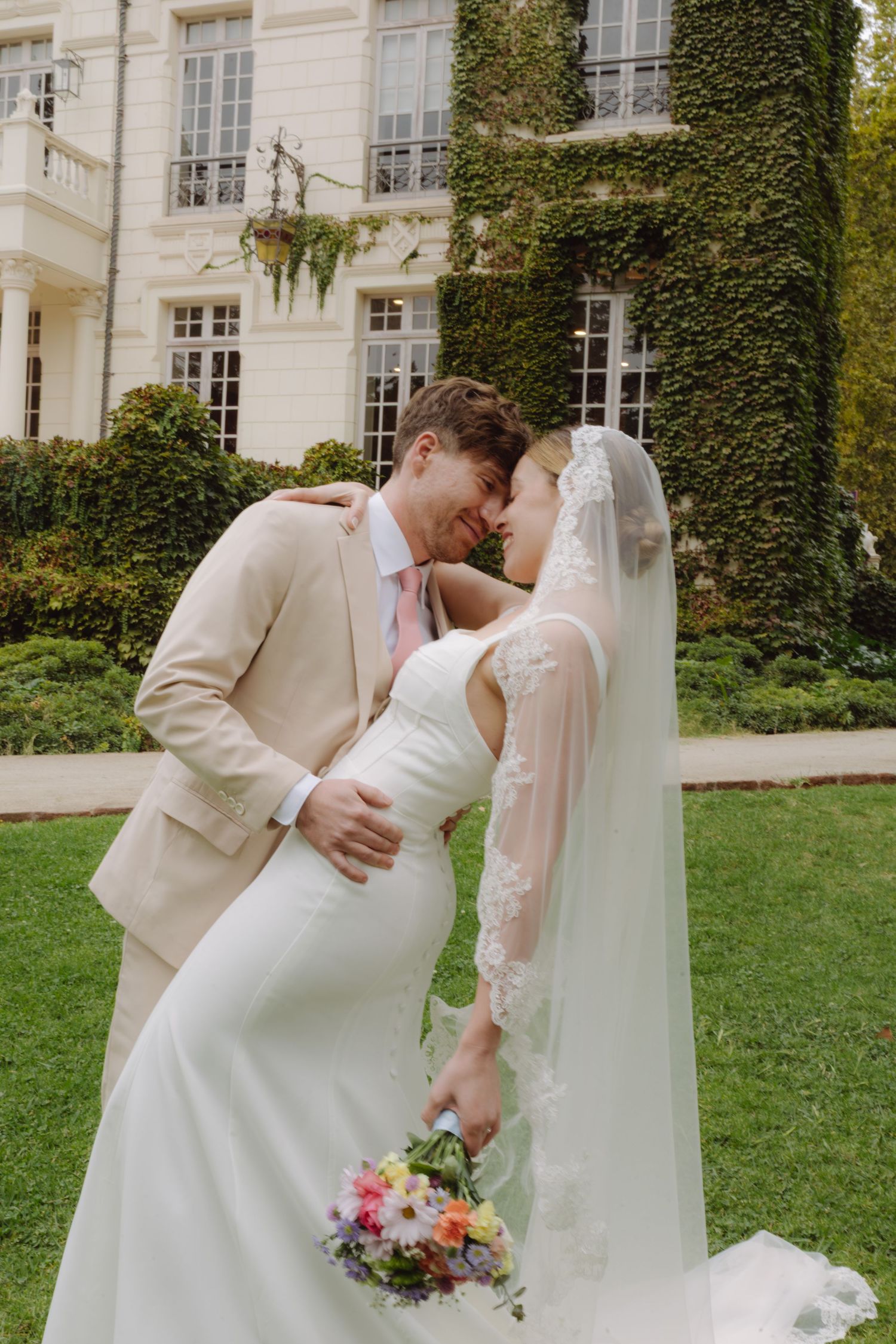 Newlyweds share an intimate nose-to-nose moment outside ivy-covered mansion, bride holding vibrant mixed flower bouquet with lace veil.
