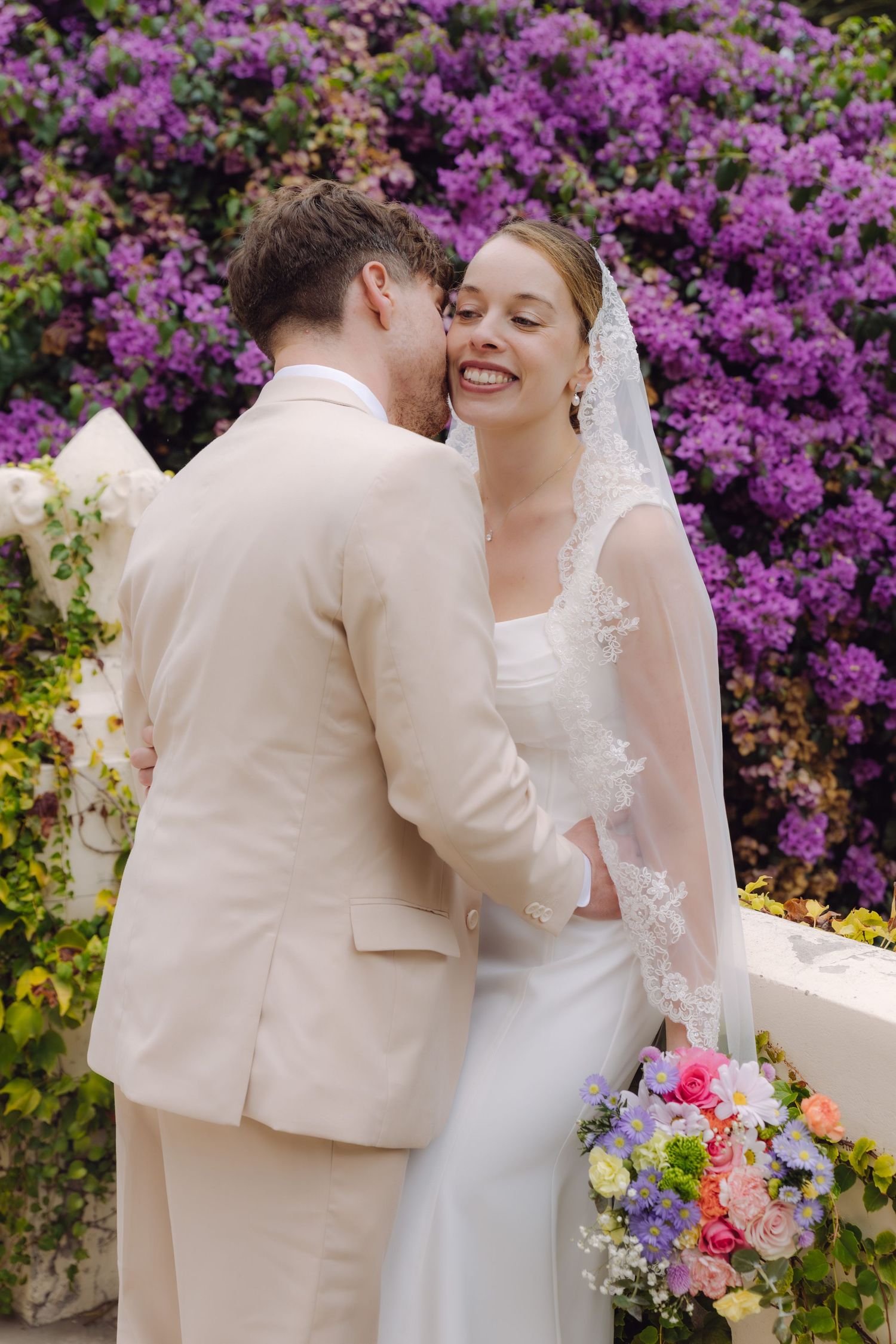 Smiling bride in white gown and lace veil leans into groom as he whispers, surrounded by purple bougainvillea blooms.
