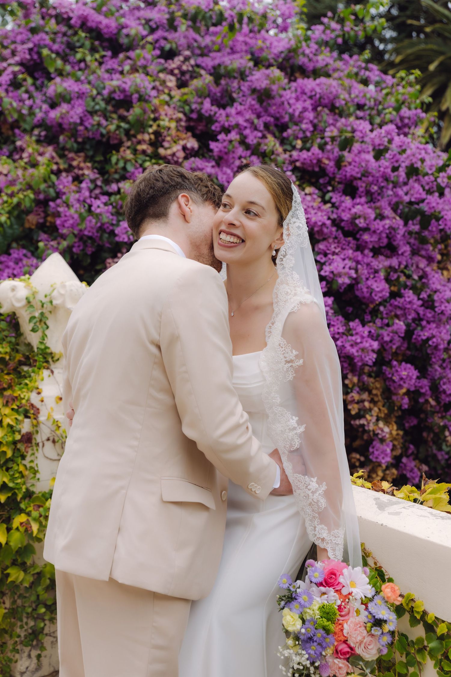 Joyful bride laughs as groom kisses her cheek against a stunning purple flowering bougainvillea backdrop, holding colorful bouquet.