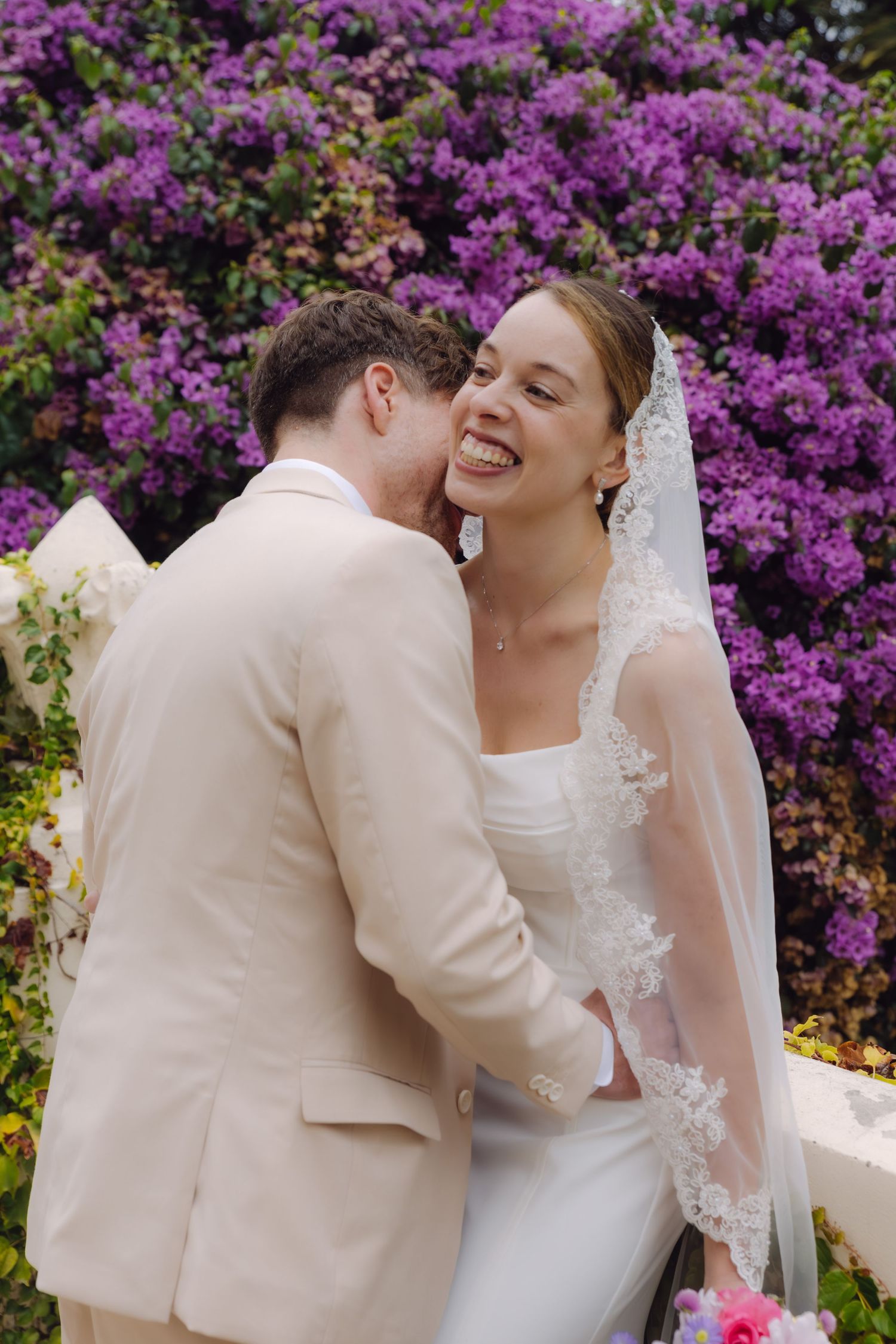 Bride laughing with eyes closed as groom nuzzles her cheek, standing before vibrant purple bougainvillea flowers outdoors.