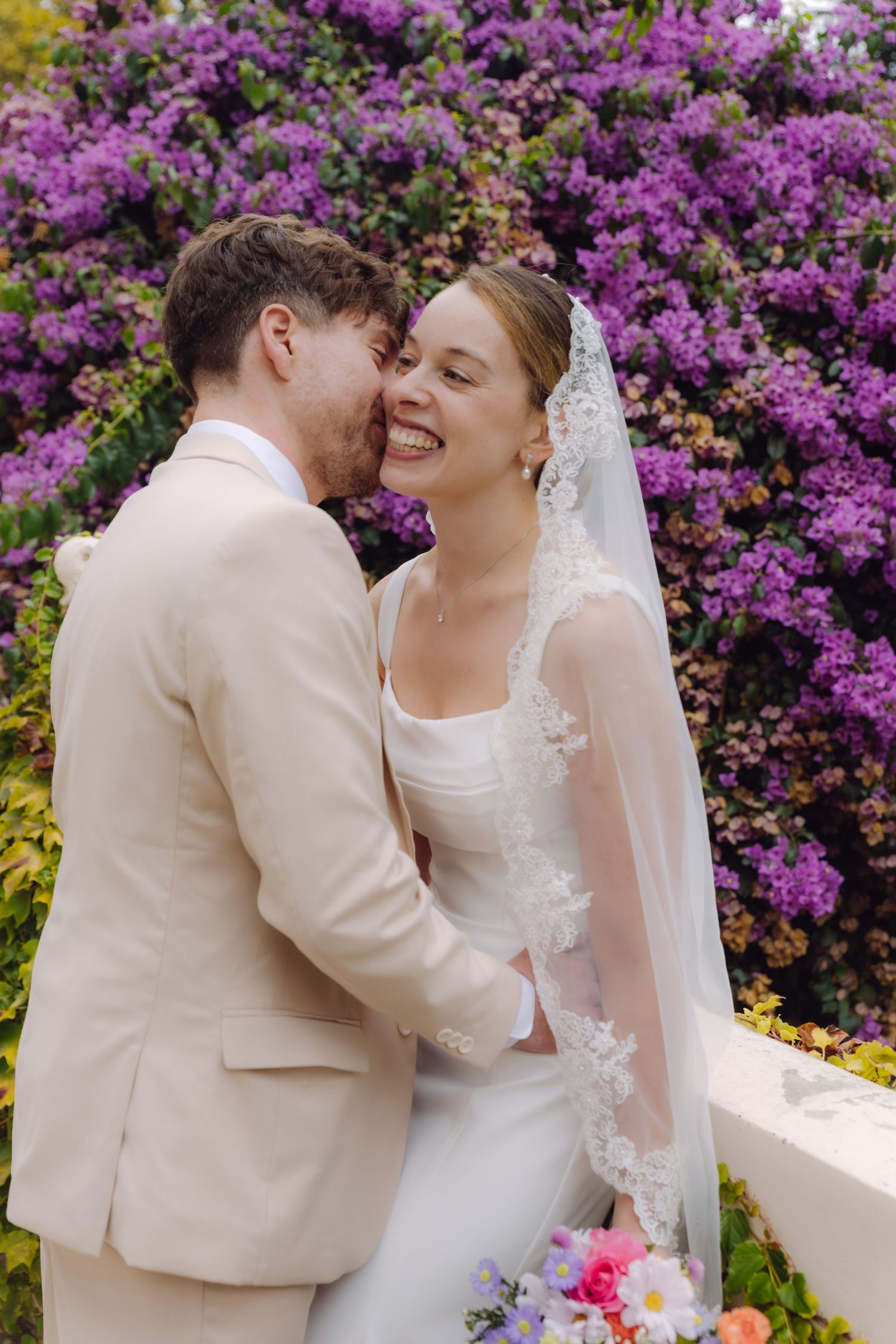 Smiling bride in lace veil and white gown embraces groom in beige suit before vibrant purple bougainvillea.