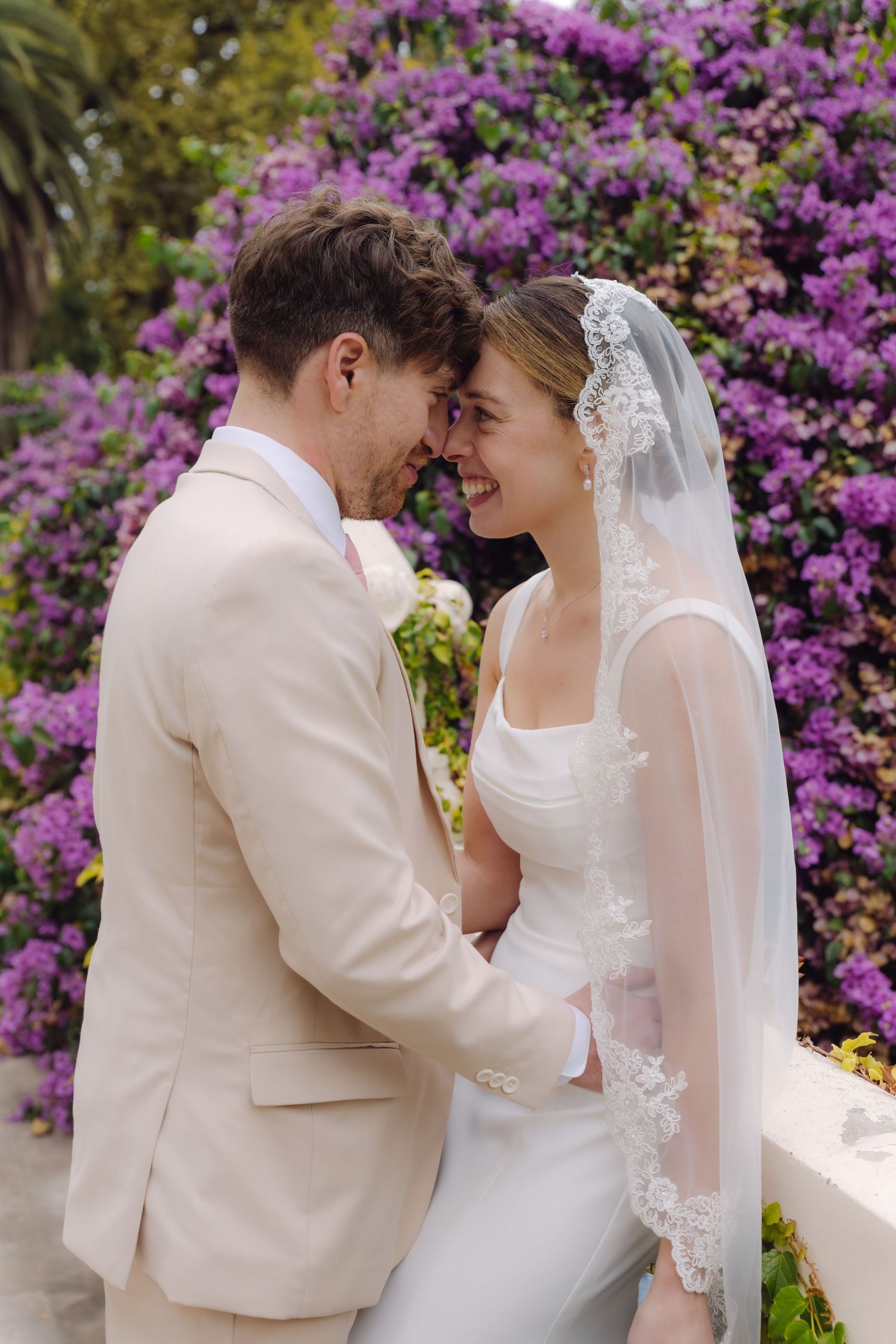 Bride and groom touch foreheads and smile tenderly in front of lush purple flowering bougainvillea backdrop.