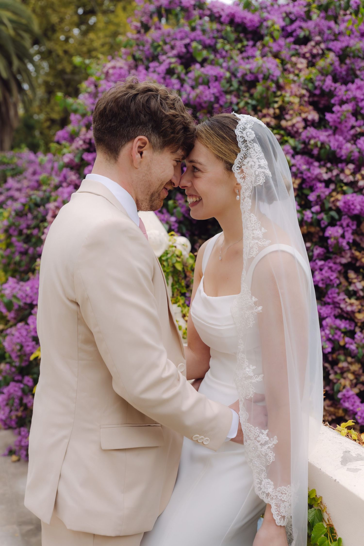 Bride in lace veil and groom in beige suit share an intimate forehead touch before purple blooming flowers.