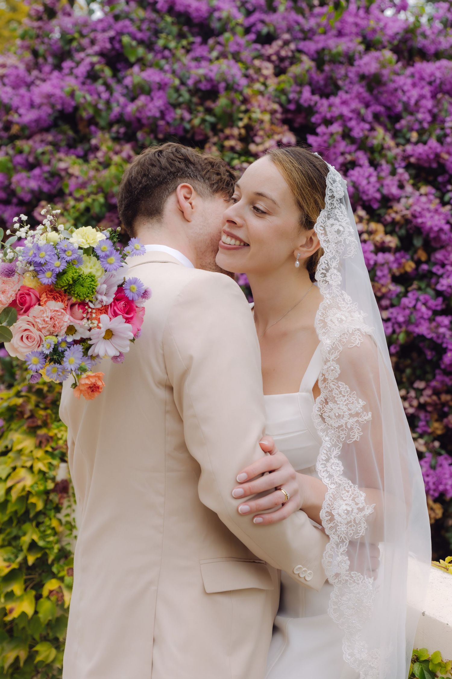 Laughing bride in lace veil embraces groom holding colorful bouquet in front of purple bougainvillea wall.