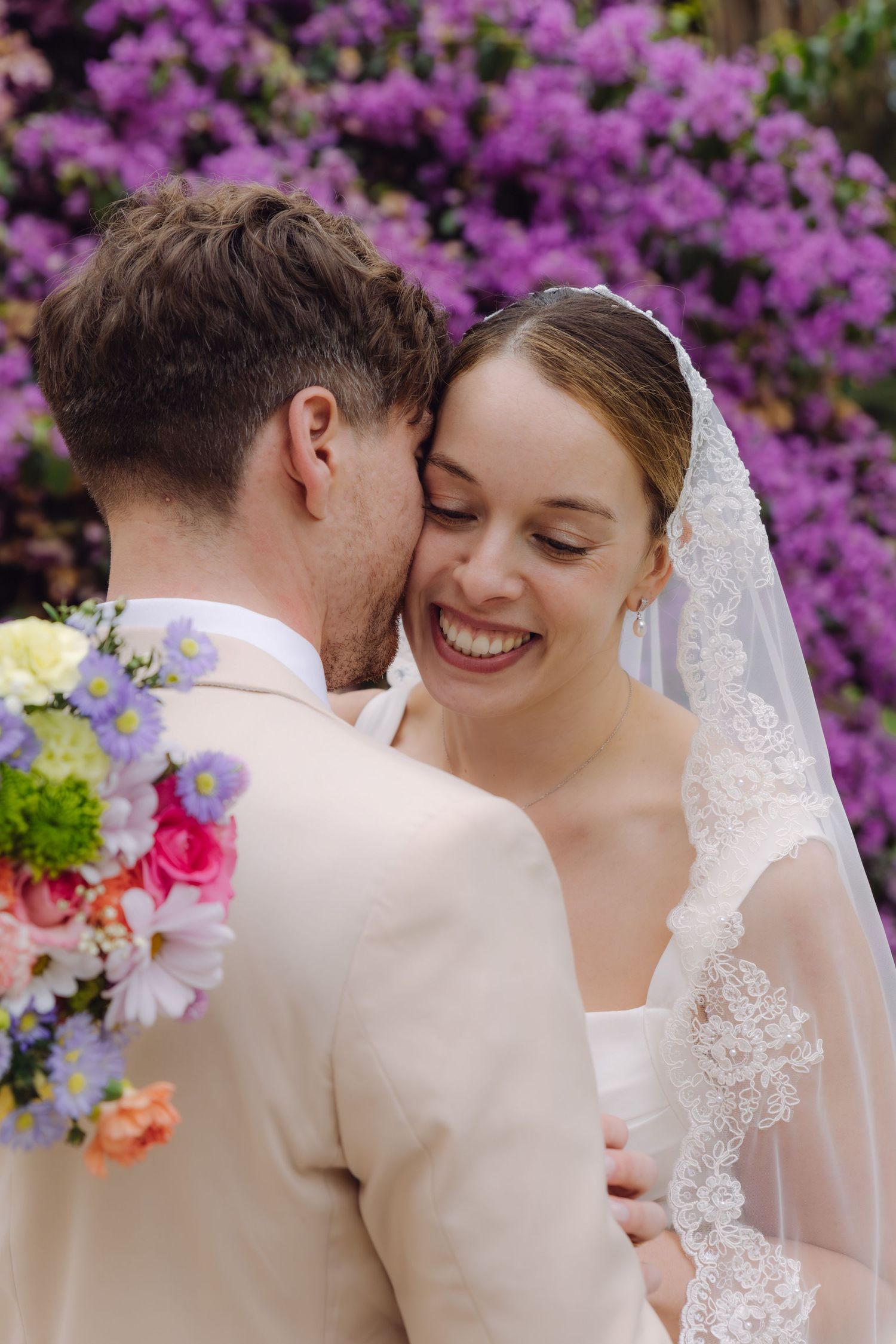 Bride in lace veil smiles joyfully as groom whispers to her, holding a vibrant mixed flower bouquet.