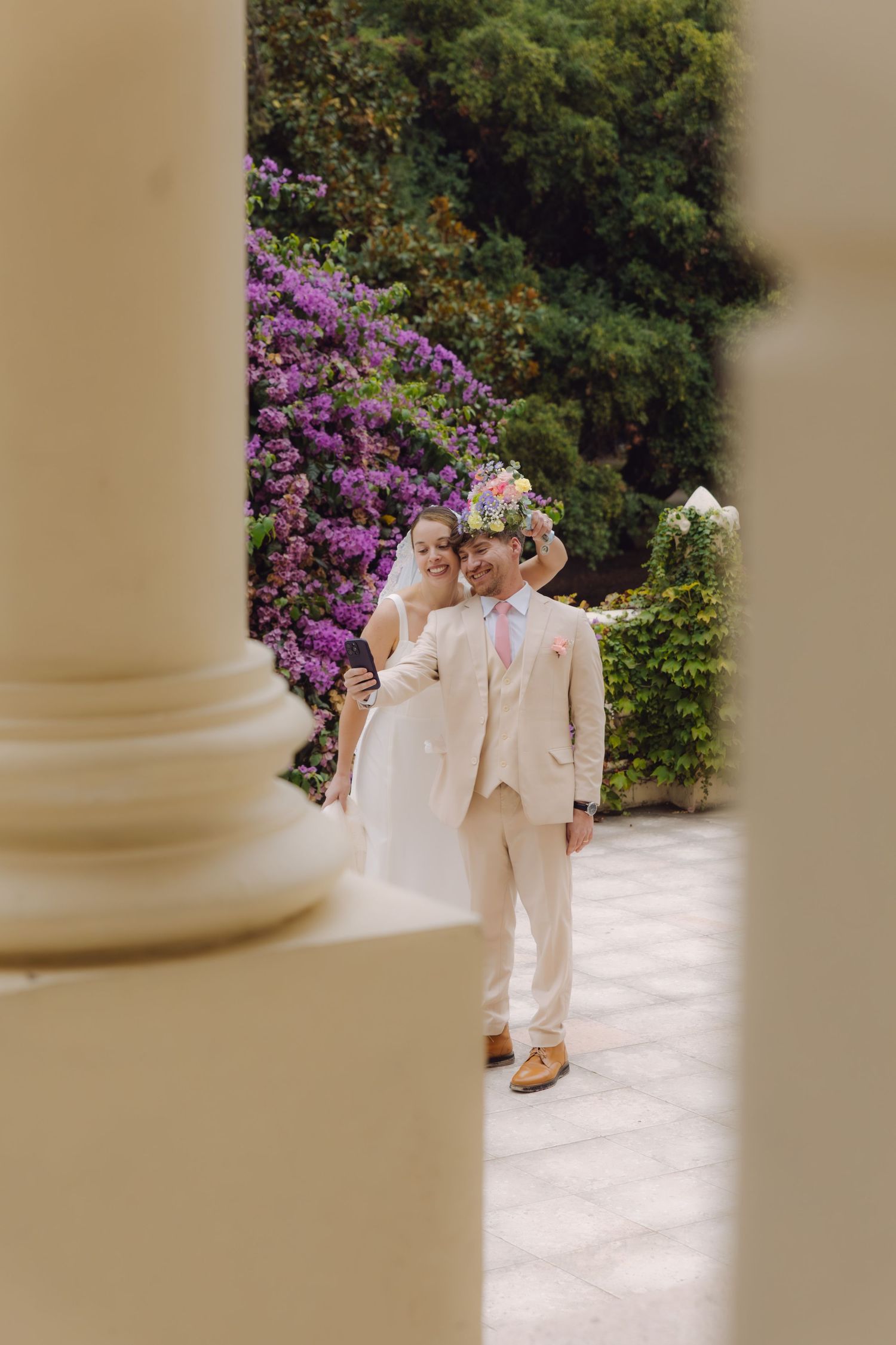 Bride and groom share a joyful moment framed by white columns with purple bougainvillea in the background.
