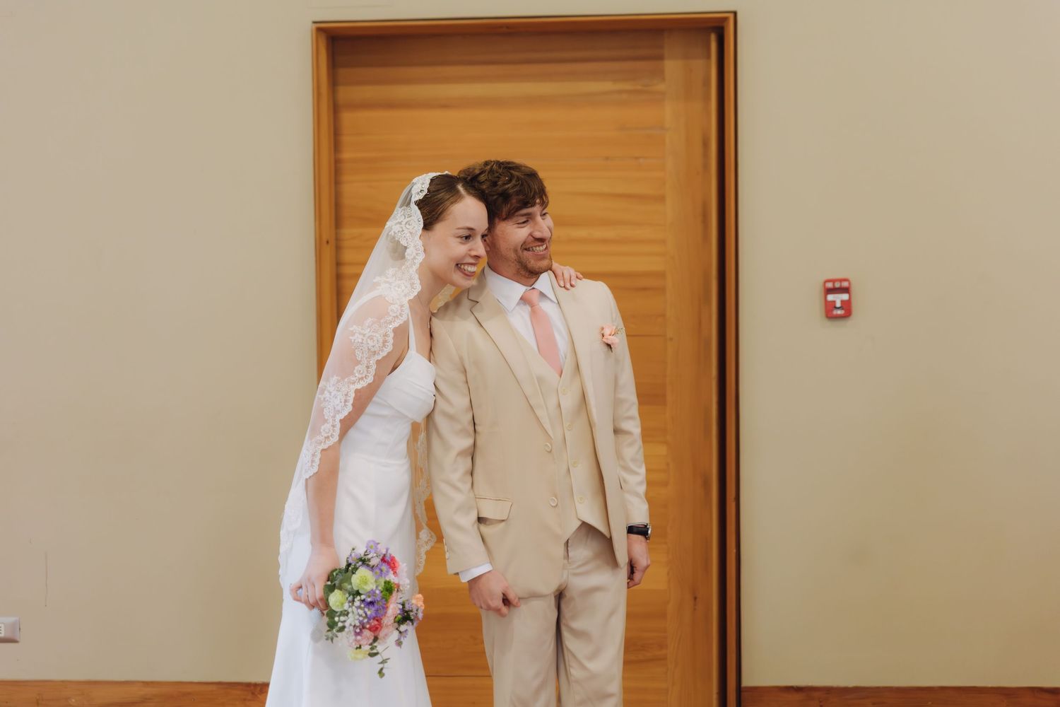 Bride in lace veil and groom in beige suit laugh together while holding a colorful bouquet indoors.