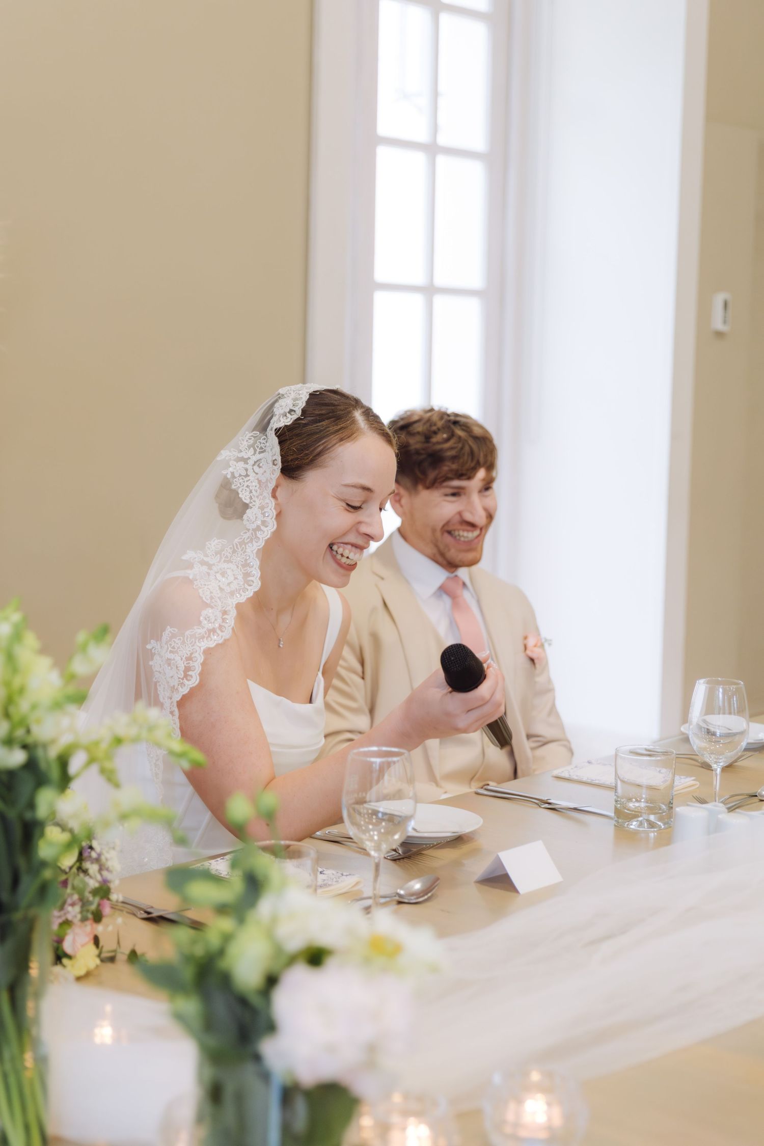 Bride laughs while speaking into a microphone at the wedding reception table beside her smiling groom.