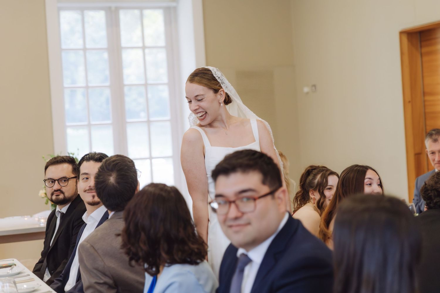 Radiant bride in white gown and veil smiles warmly while greeting seated wedding reception guests.