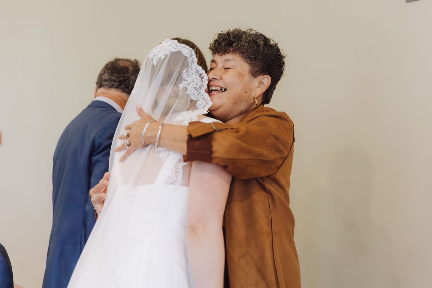 Joyful wedding guest in brown jacket hugs the bride in a white gown and veil at the reception.