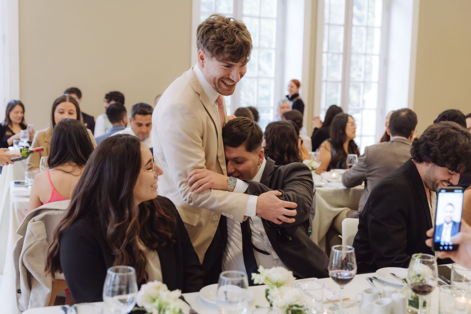 Smiling young man in beige suit hugs a seated guest at a wedding reception dinner.