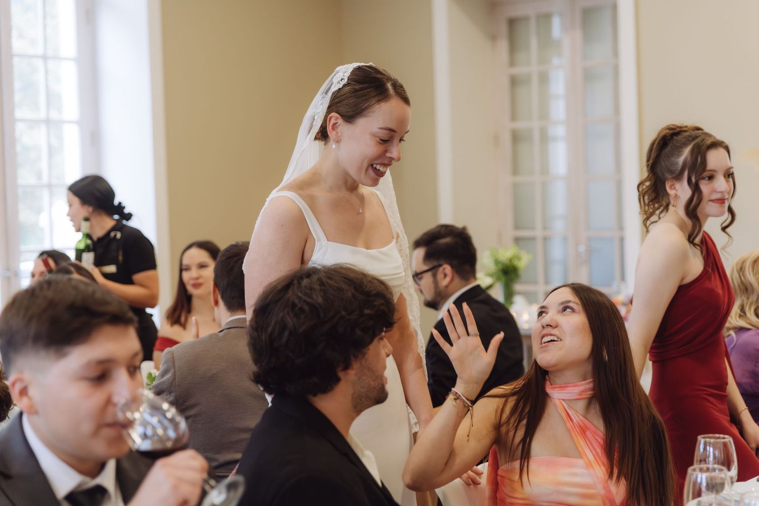 Radiant bride in white veil greets guests at wedding reception tables with bridesmaids nearby.