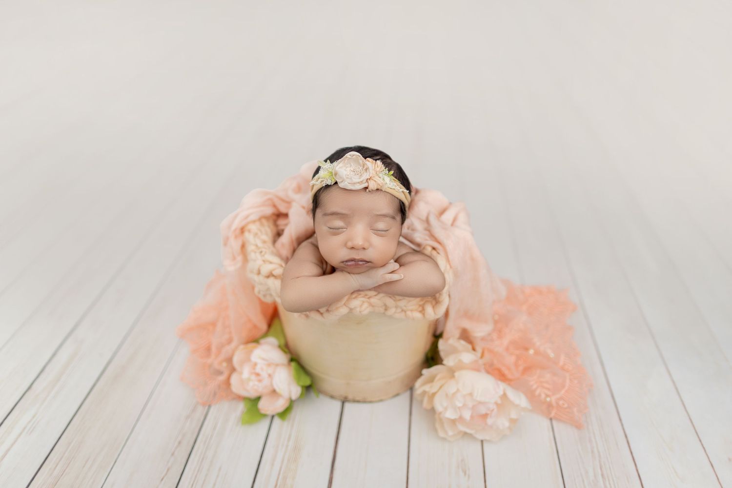 Newborn baby girl sleeping peacefully in a wooden bucket with peach flowers and floral headband on white wood floor.