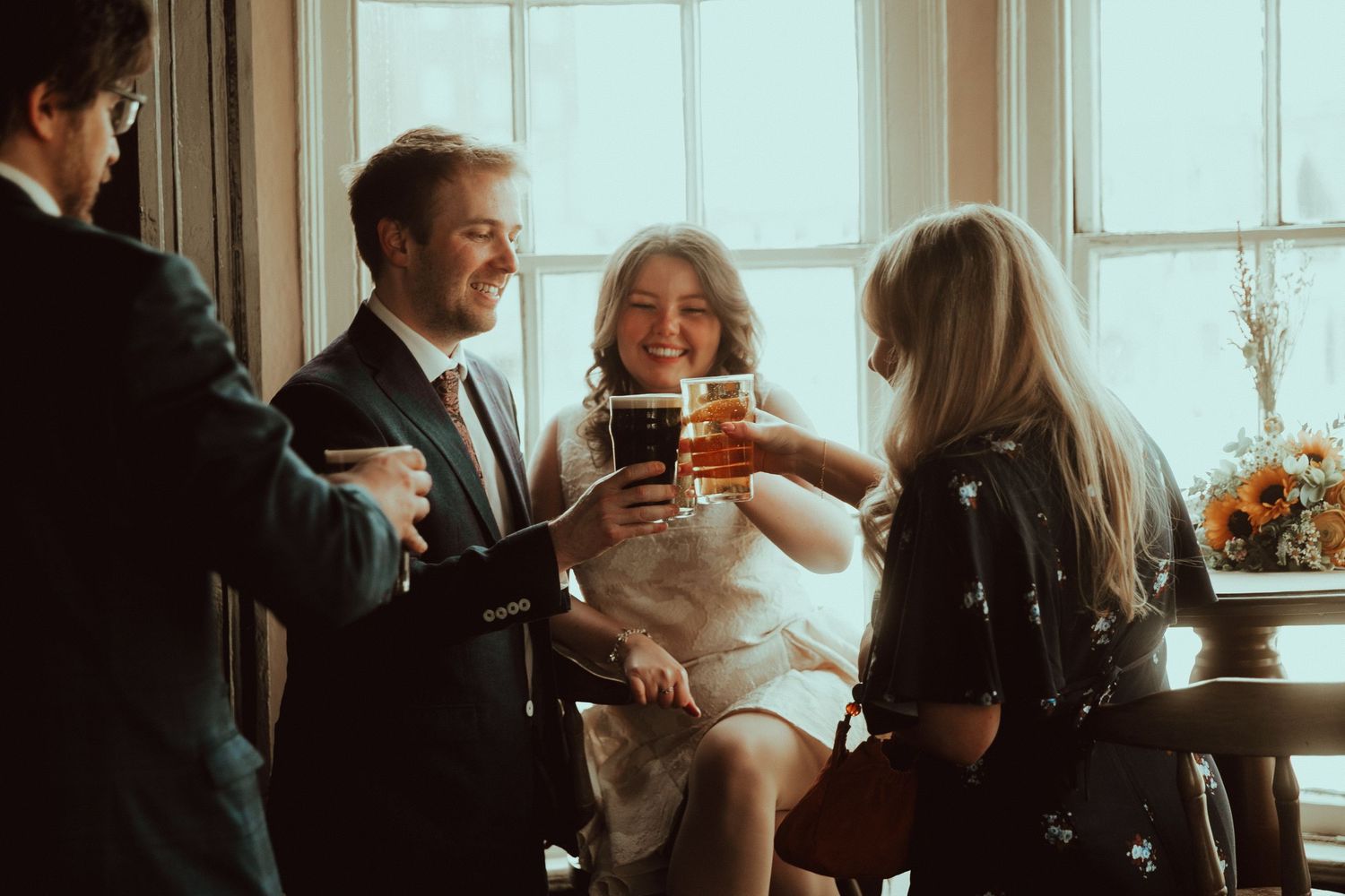 Elegantly dressed group of friends toasting with drinks at a bright window during a formal celebration.