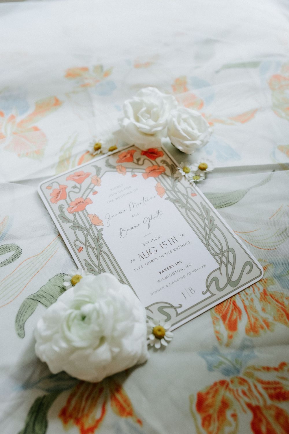 Elegant wedding invitation with Art Nouveau floral design, surrounded by white roses and daisies on a floral fabric background.