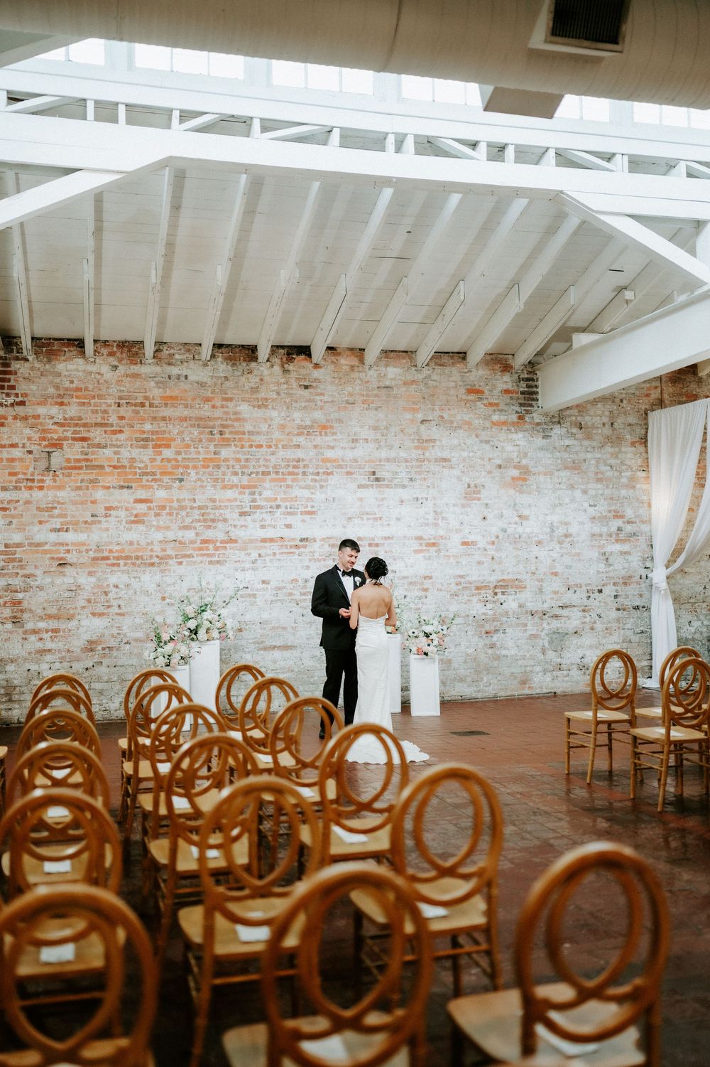 Bride and groom share first dance in elegant brick-walled venue with white rafters and gold chiavari chairs.
