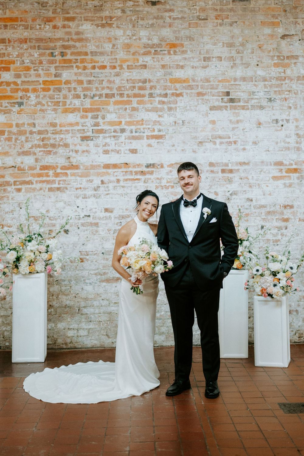 Bride in white gown and groom in black tuxedo pose before a rustic brick wall with floral arrangements.