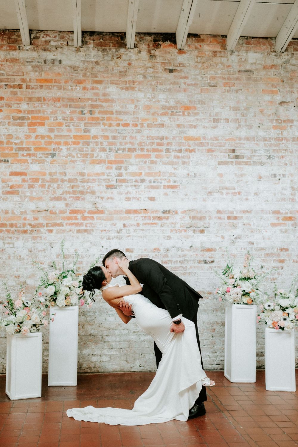 Bride and groom share a romantic dip kiss in front of a rustic brick wall flanked by white pedestals with floral arrangements.