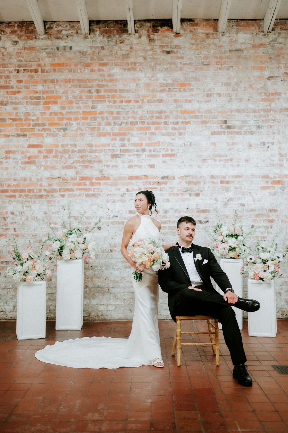 Bride in white halter gown and groom in black tuxedo pose before rustic brick wall with white pedestal floral arrangements.