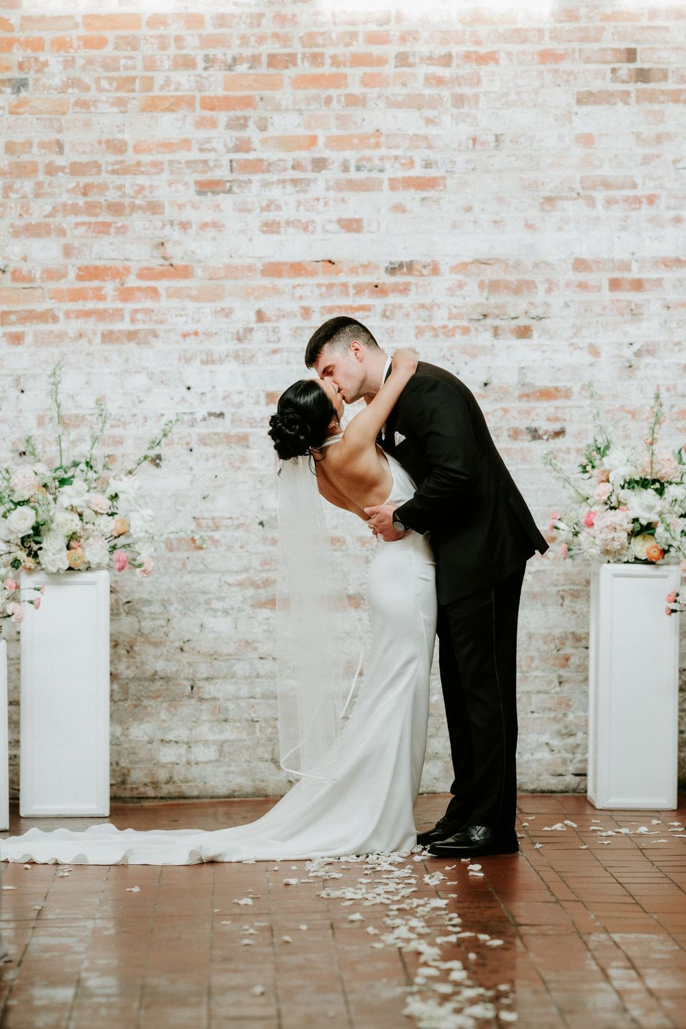Bride and groom share first kiss at wedding ceremony in front of brick wall with floral arrangements.
