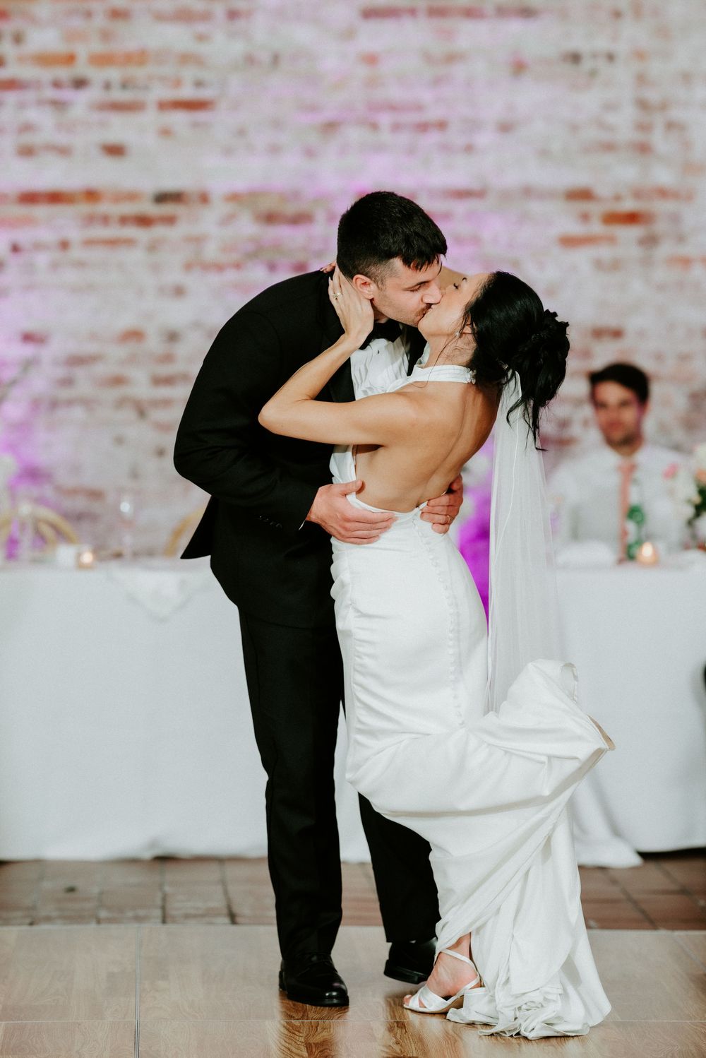 Bride and groom share a romantic dip kiss on the dance floor at their wedding reception with brick wall backdrop.