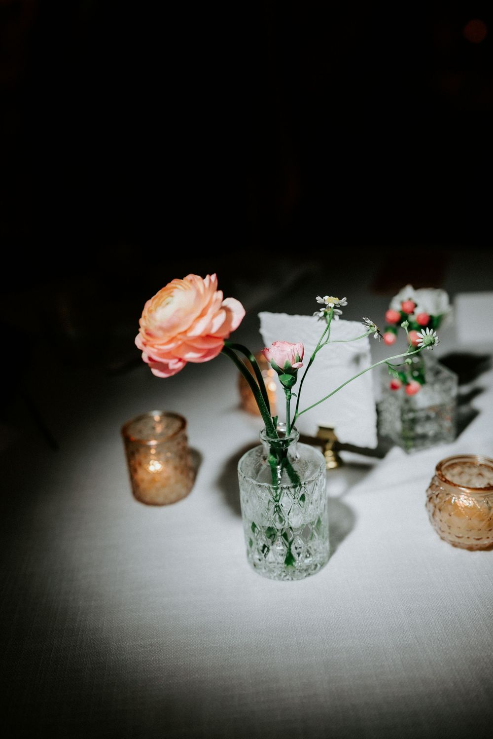 Elegant wedding tablescape featuring a peach ranunculus in a crystal bud vase with amber votives on white linen.