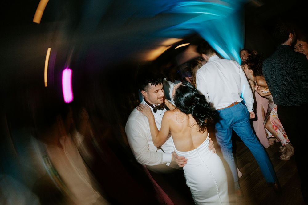 Bride and groom share a dance with guests at a vibrant wedding reception with colorful lighting.