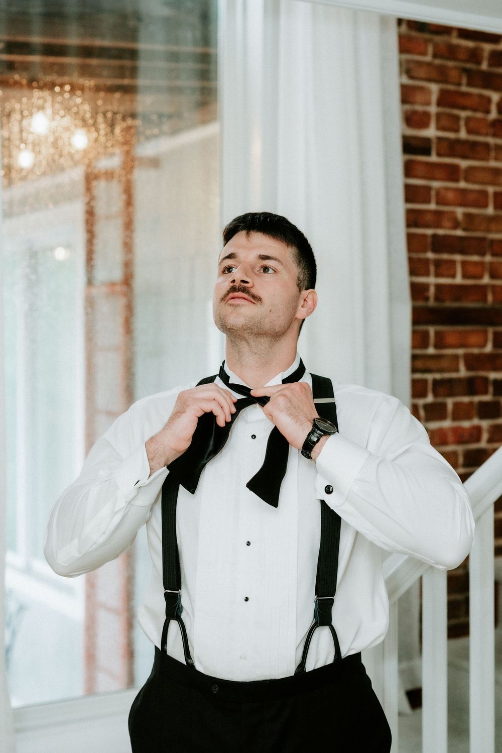 A groom in a white tuxedo shirt adjusts his black bow tie while wearing suspenders in an elegant venue.