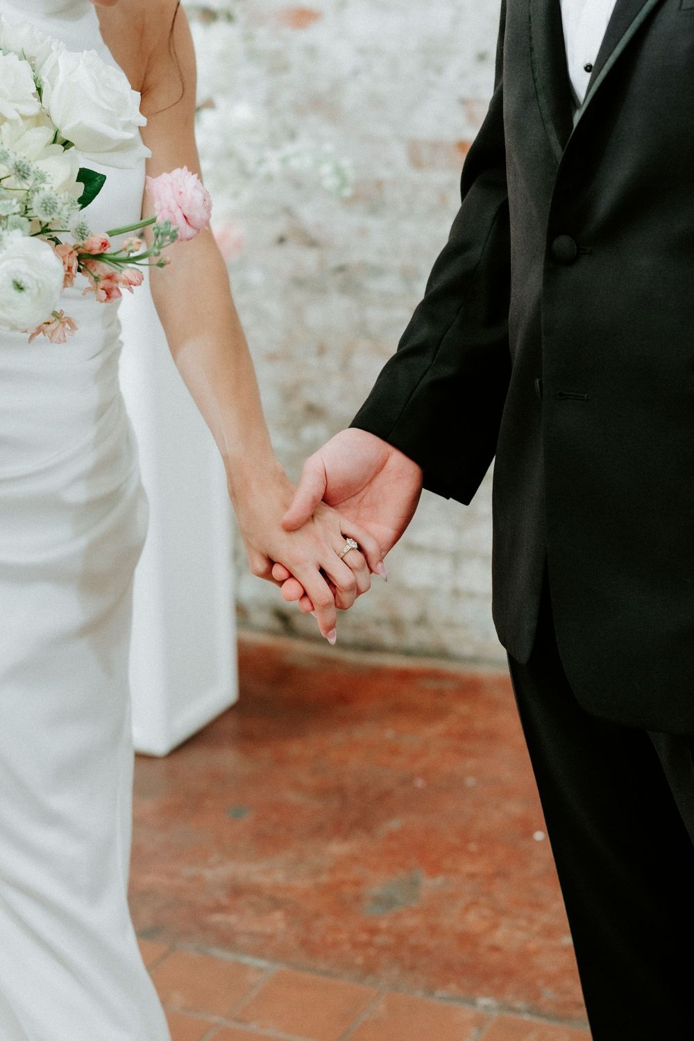 Bride and groom holding hands, bride wearing wedding ring and holding white floral bouquet, groom in black tuxedo.