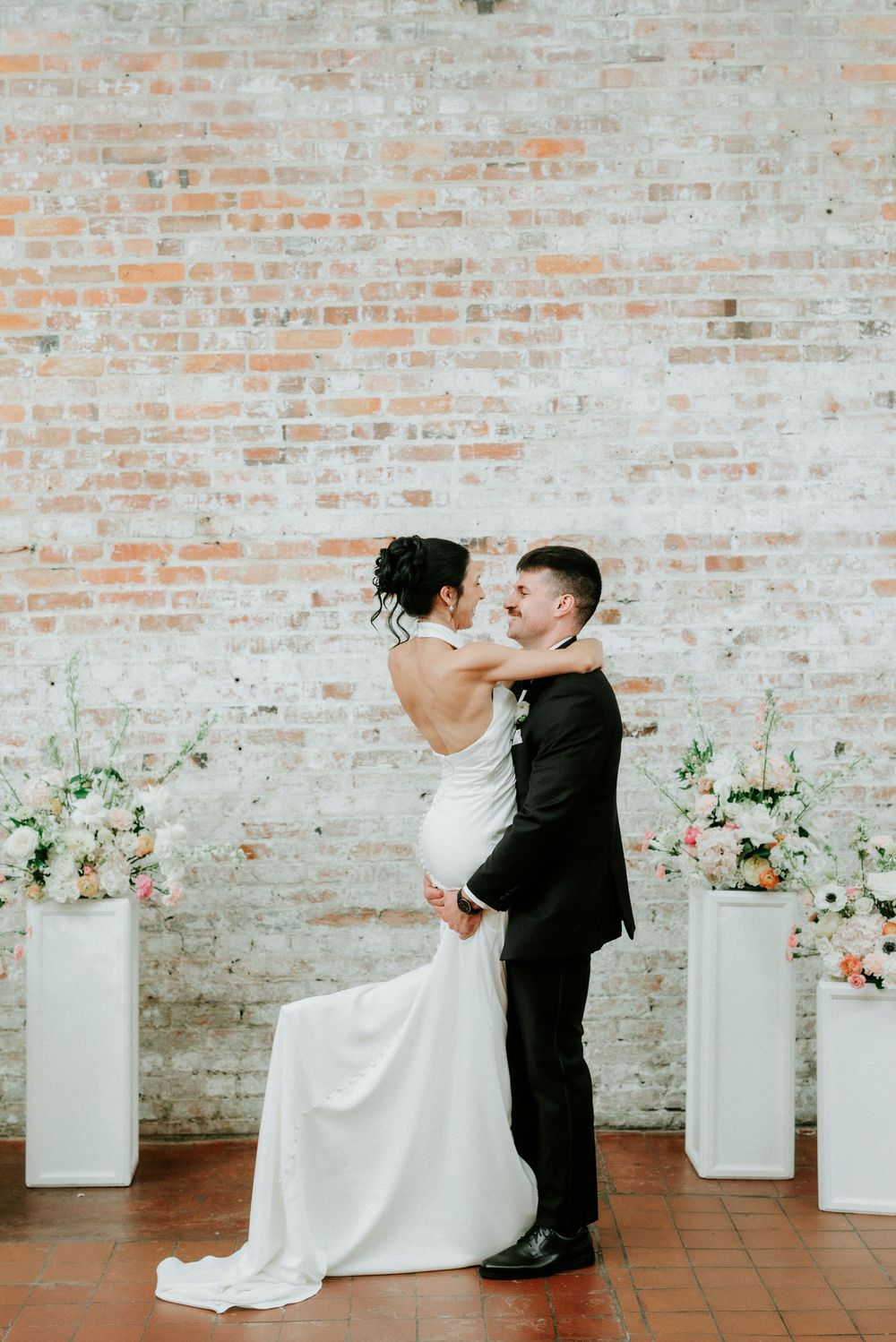 Bride and groom embrace and smile at each other during wedding ceremony in front of a rustic brick wall with floral pedestals.