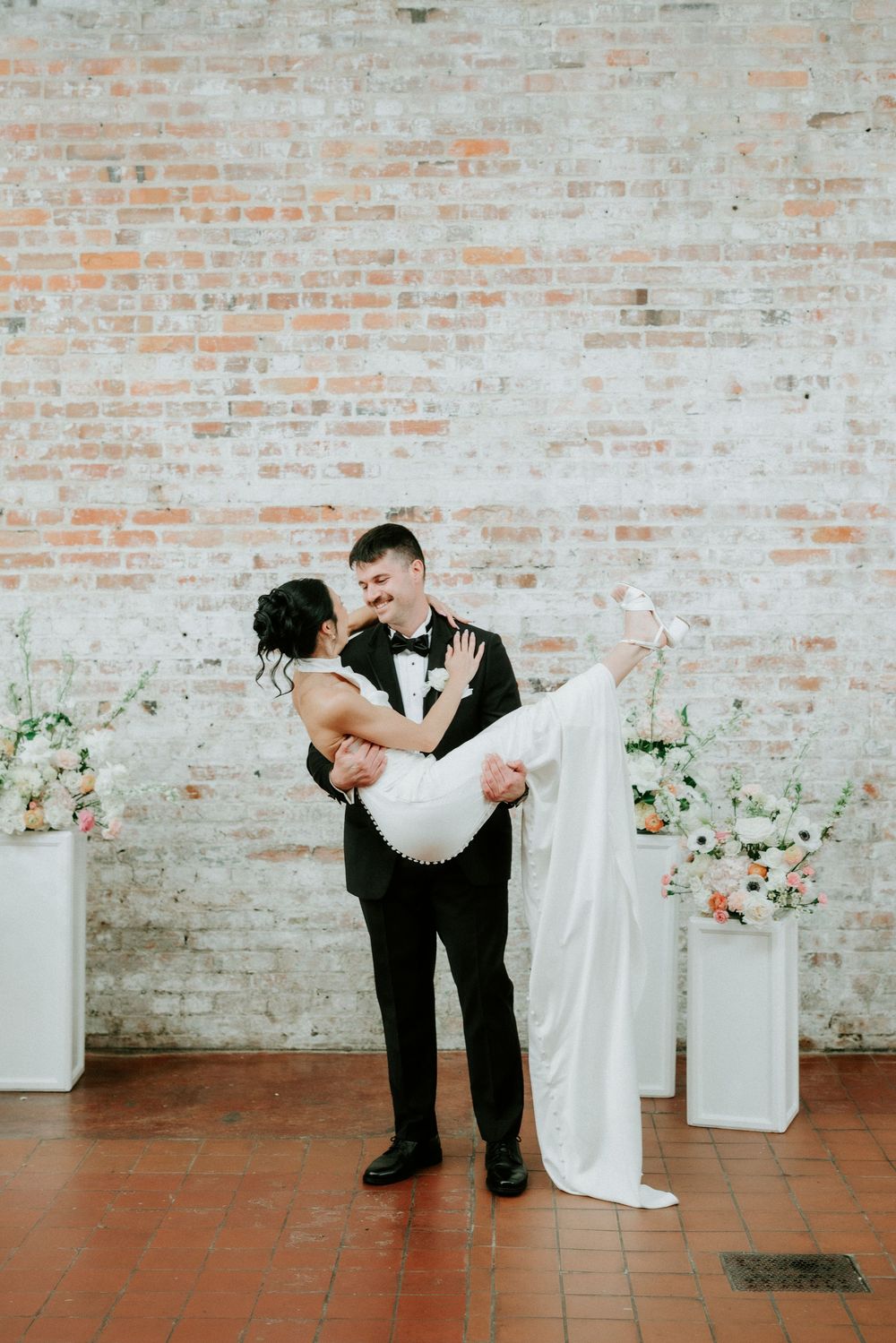 Groom in black tuxedo lifts bride in white gown, posing before rustic brick wall with floral arrangements.