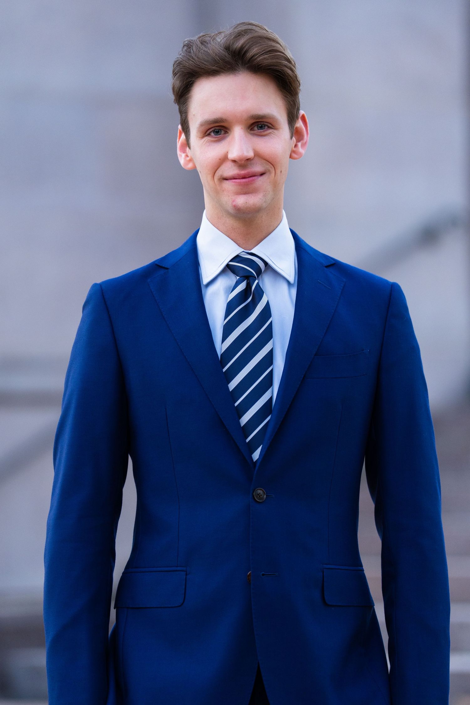 Young professional man in navy blue suit and striped tie smiling outdoors on grey steps.