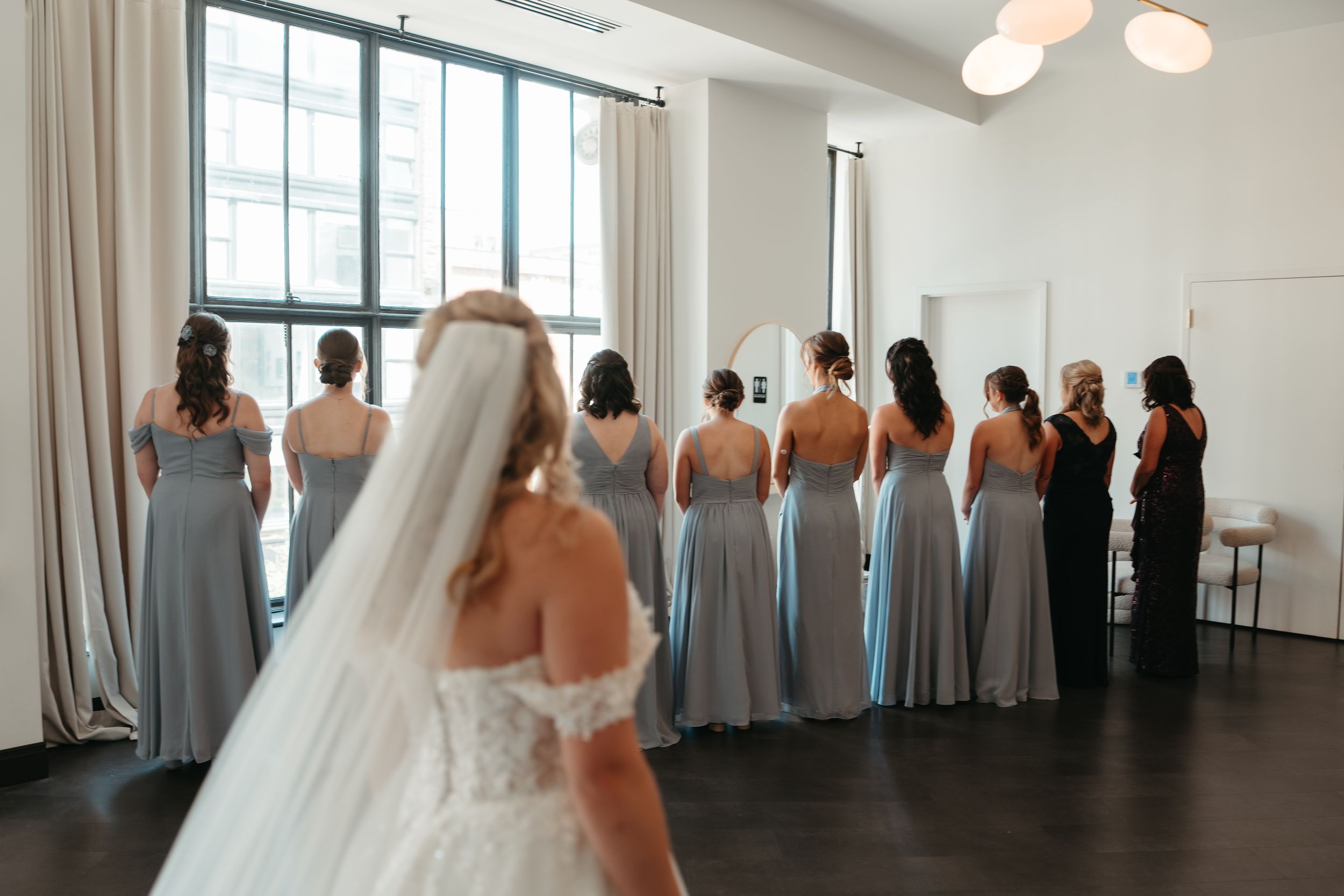 Bride in white gown and veil approaches bridesmaids in blue-gray dresses standing in a row facing away in bright room.
