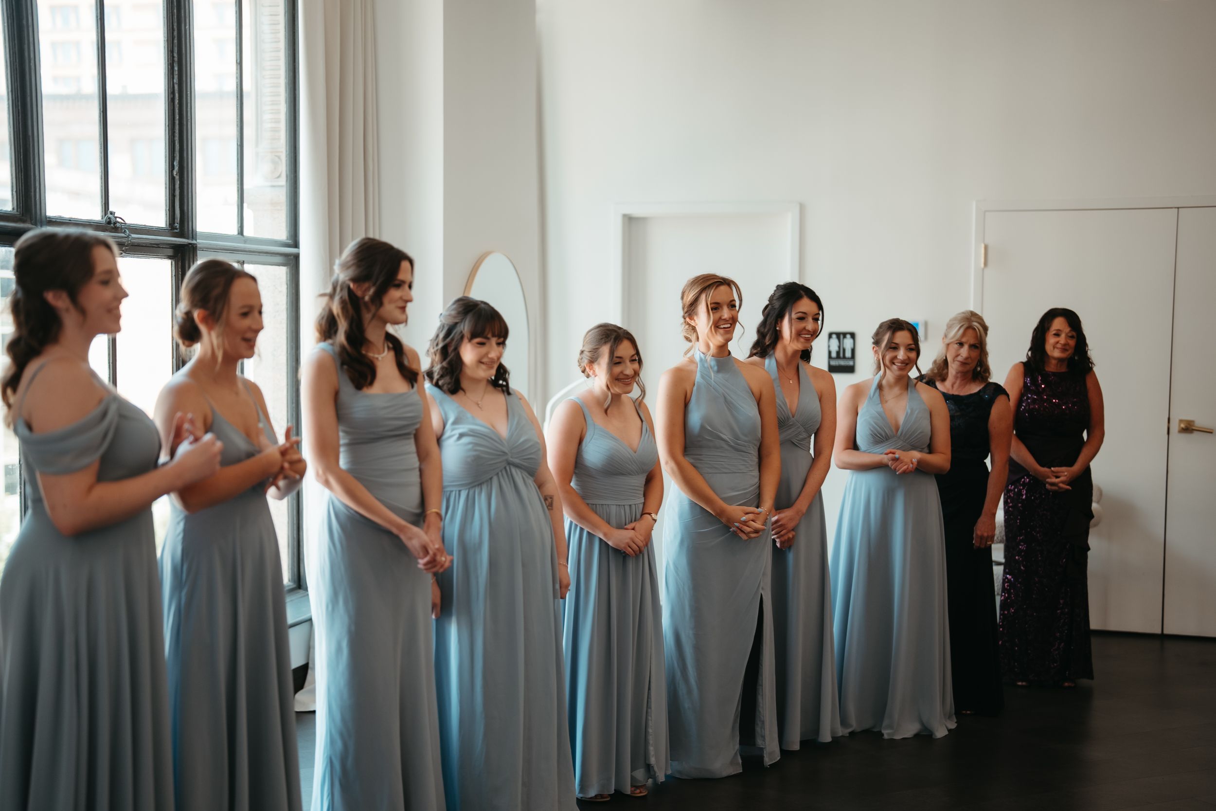 Bridesmaids in light blue floor-length gowns stand in a row during wedding preparations in a bright white room.