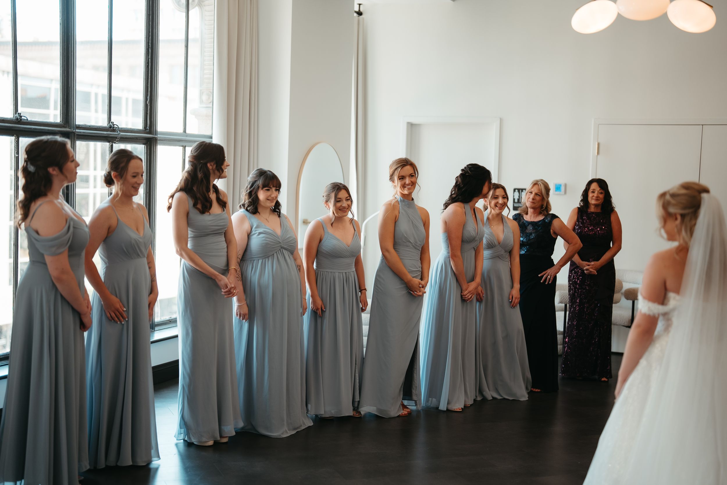 Bridesmaids in matching dusty blue gowns line up to see the bride in a bright modern wedding venue.
